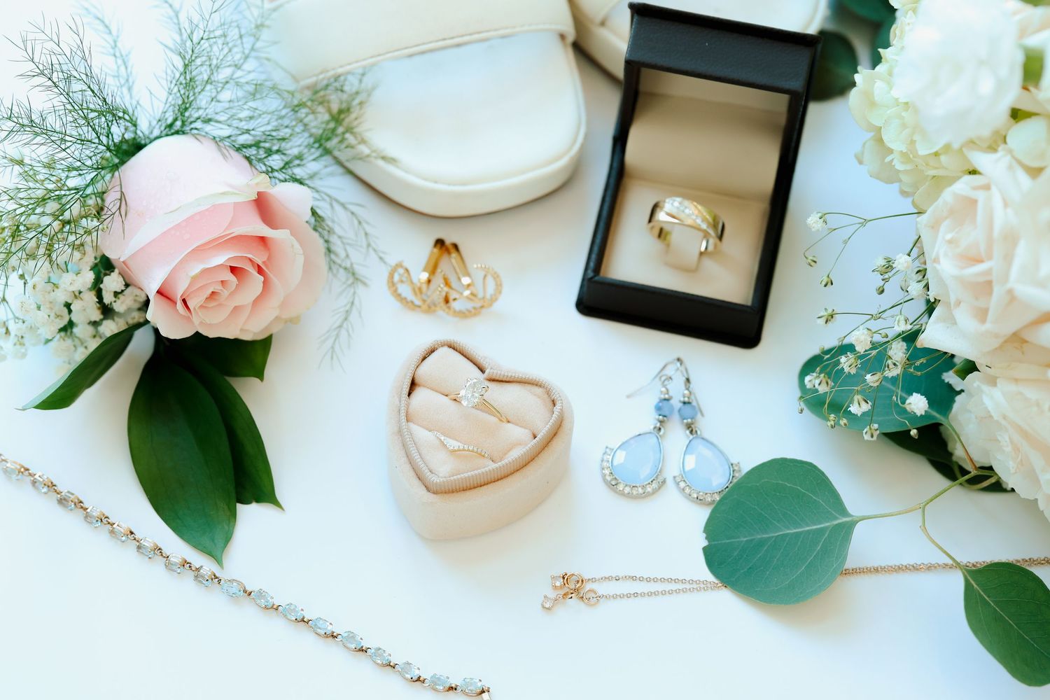 Bridal jewelry flat lay with rings, earrings, necklace, and pink rose on white surface.