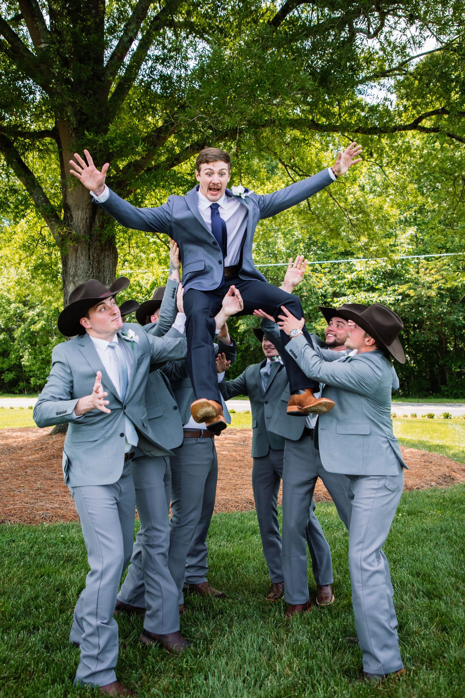 Groomsmen in gray suits and cowboy hats toss the groom into the air in a fun celebratory wedding photo.