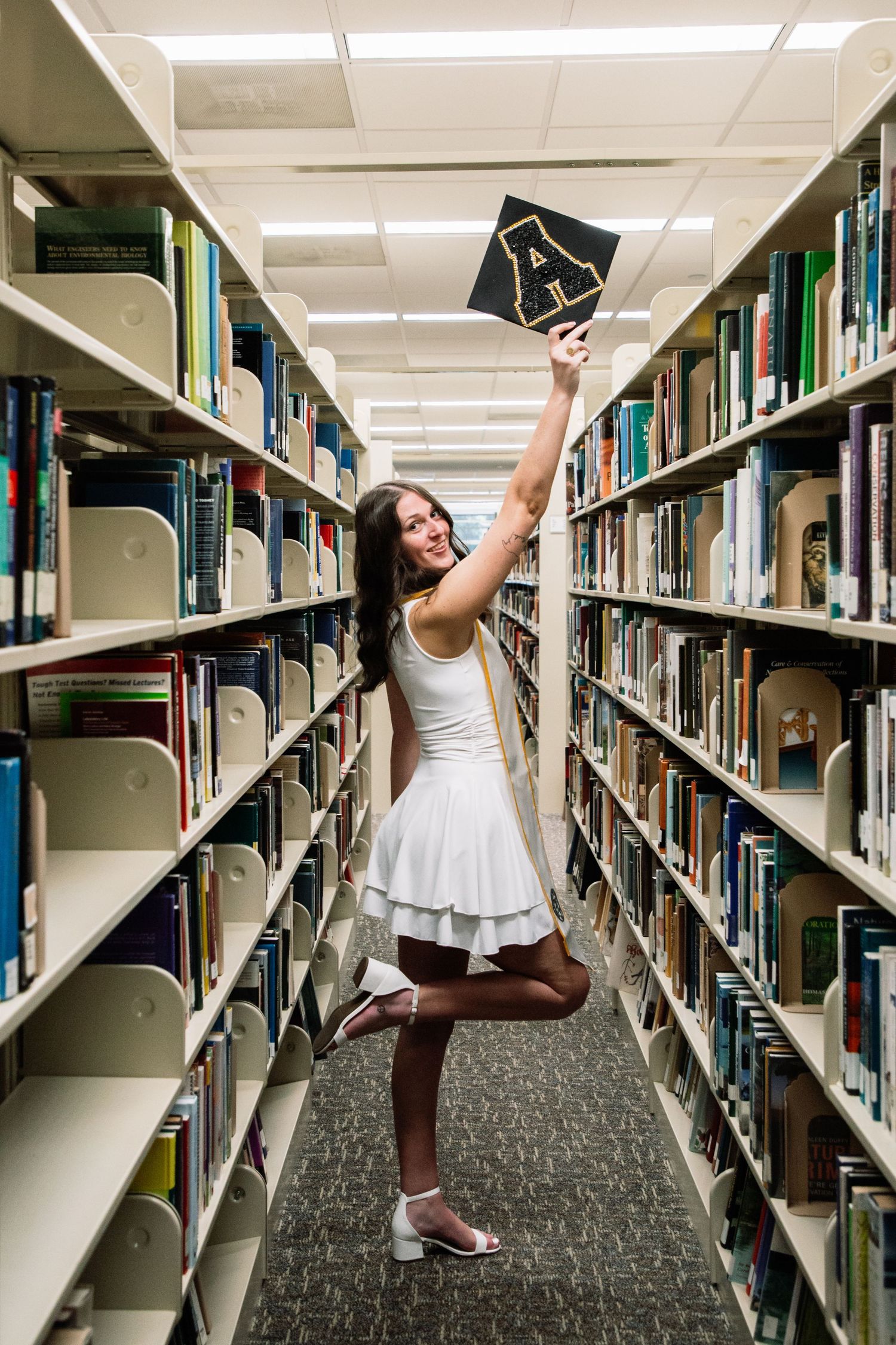 Smiling female graduate in white dress raises graduation cap in library aisle between bookshelves.