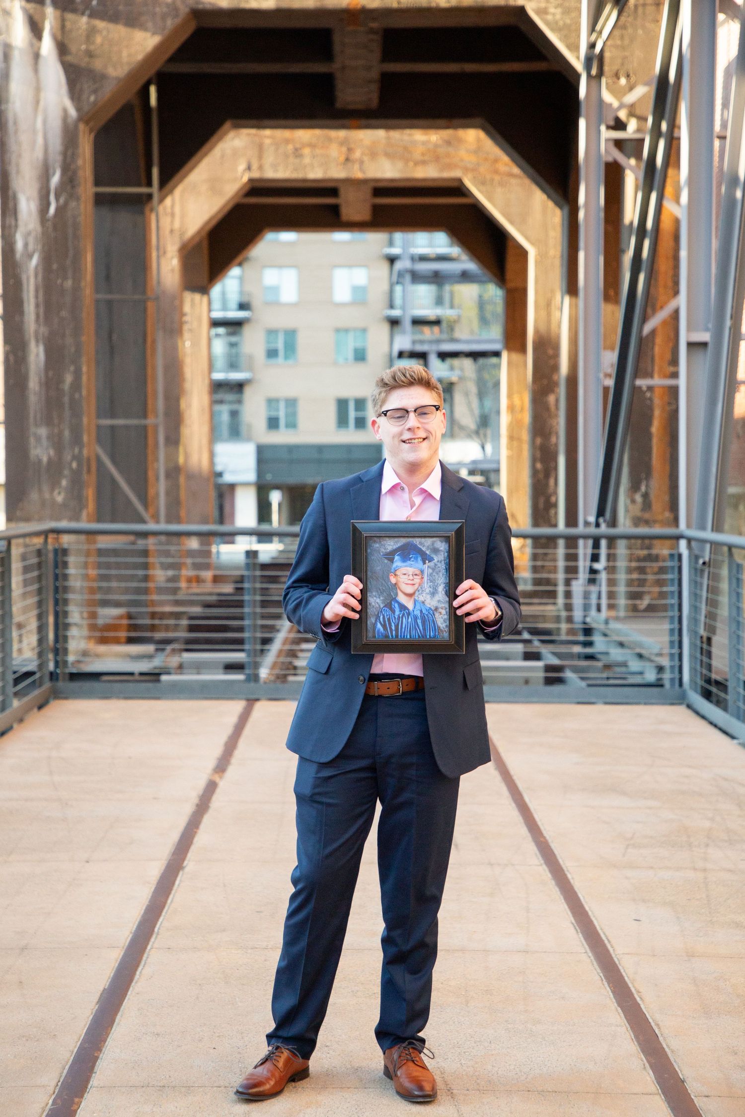 Young man in navy suit holding a framed graduation portrait on an industrial bridge.