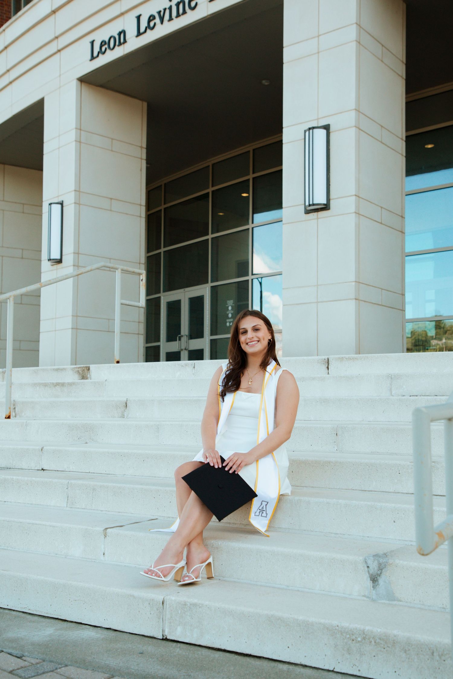 Smiling graduate in white dress sits on steps outside Leon Levine Hall holding diploma.