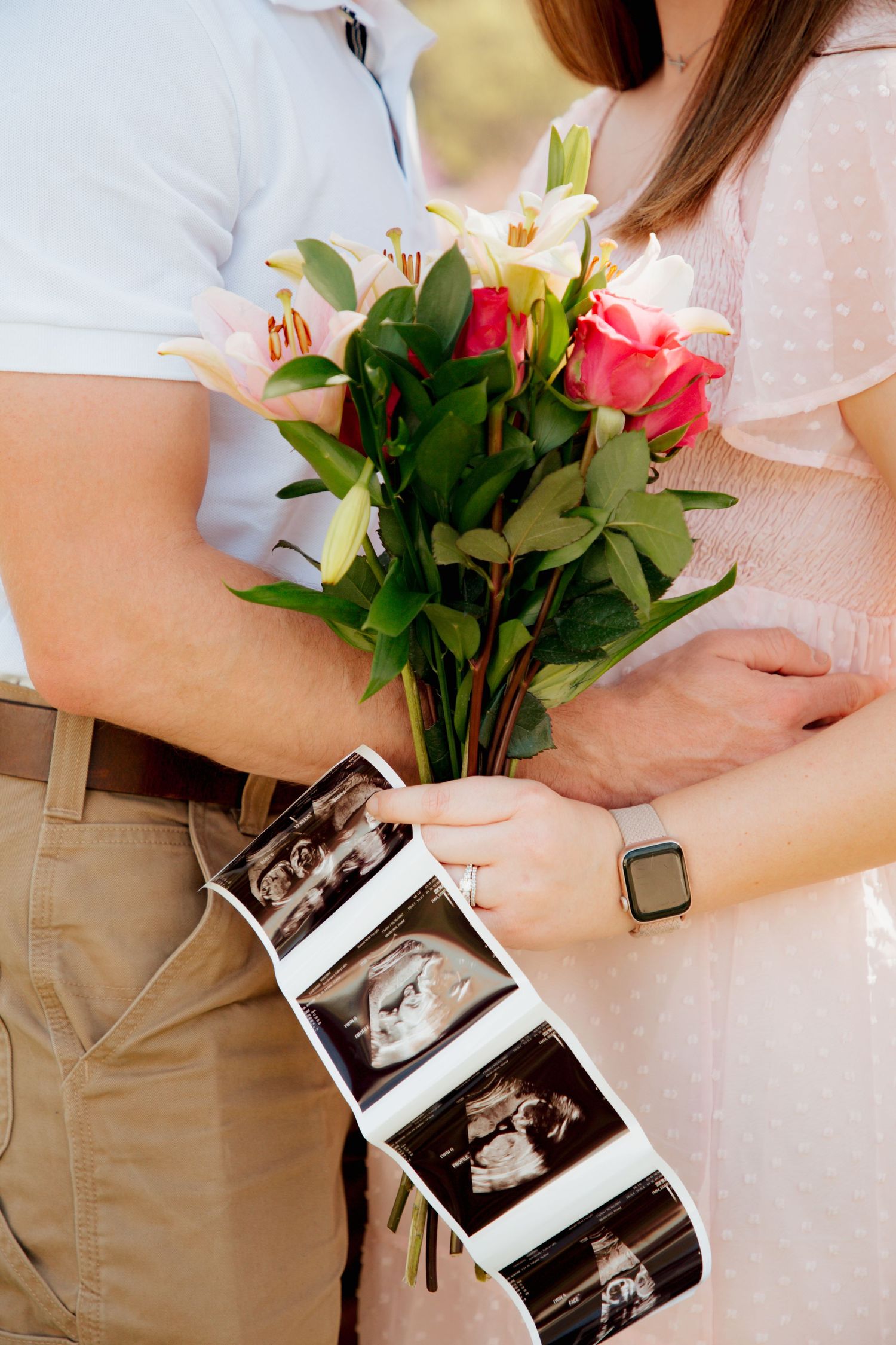 Close-up of couple holding pink flower bouquet and ultrasound photos together outdoors.