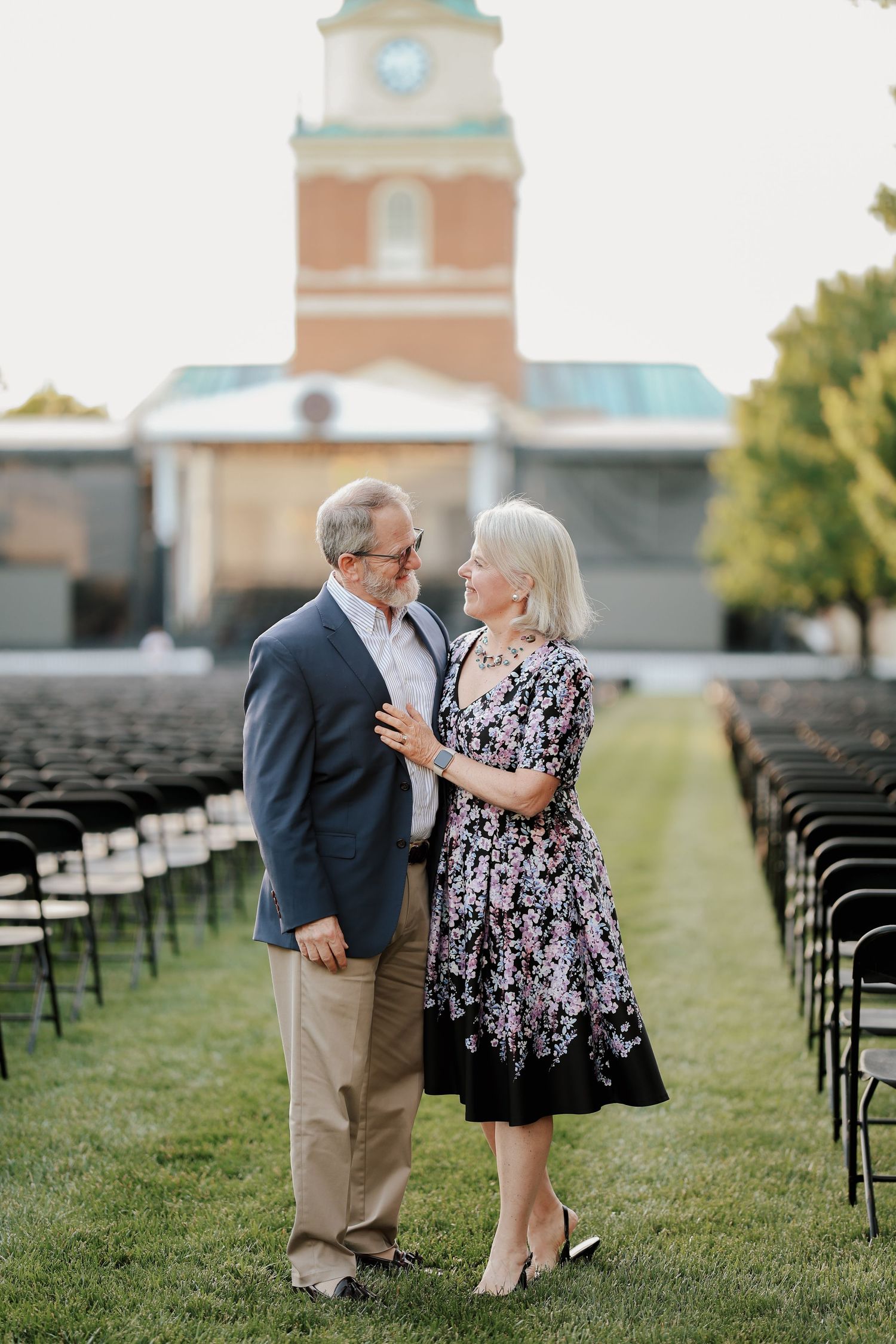 Older couple gazing at each other lovingly outdoors near rows of black chairs and brick building.