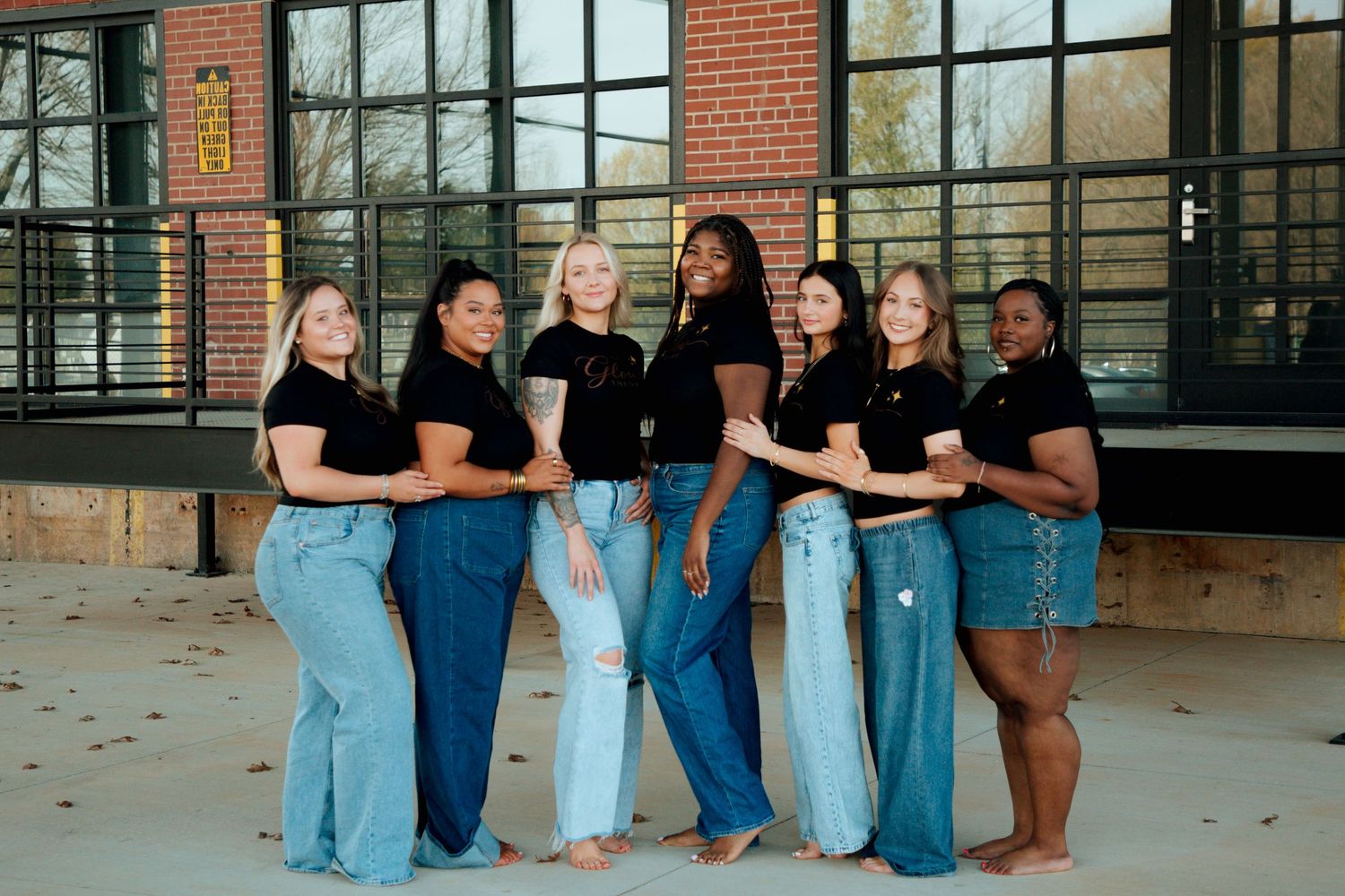 Seven diverse women in black tops and blue jeans posing together in front of large windows outdoors.