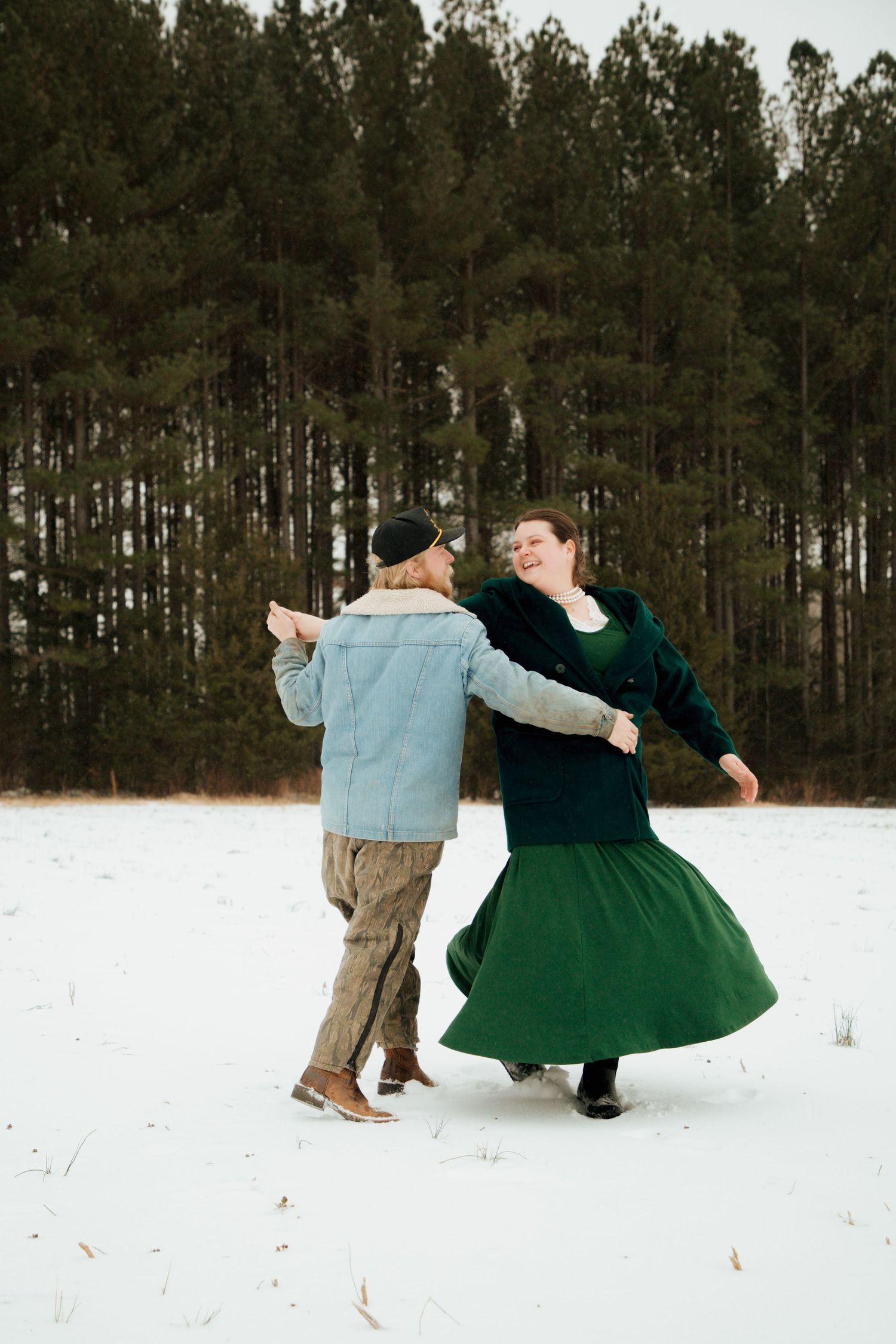Joyful couple dancing and spinning in snow-covered field with tall pine trees in background.