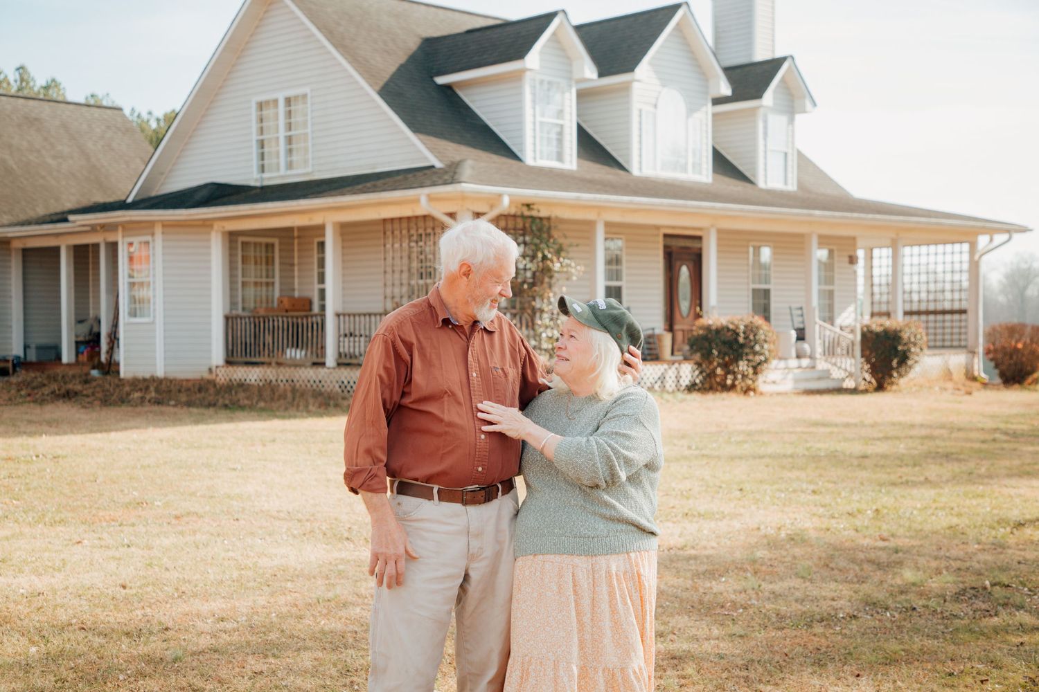 Elderly couple sharing a tender moment in front of a white farmhouse on a sunny day.