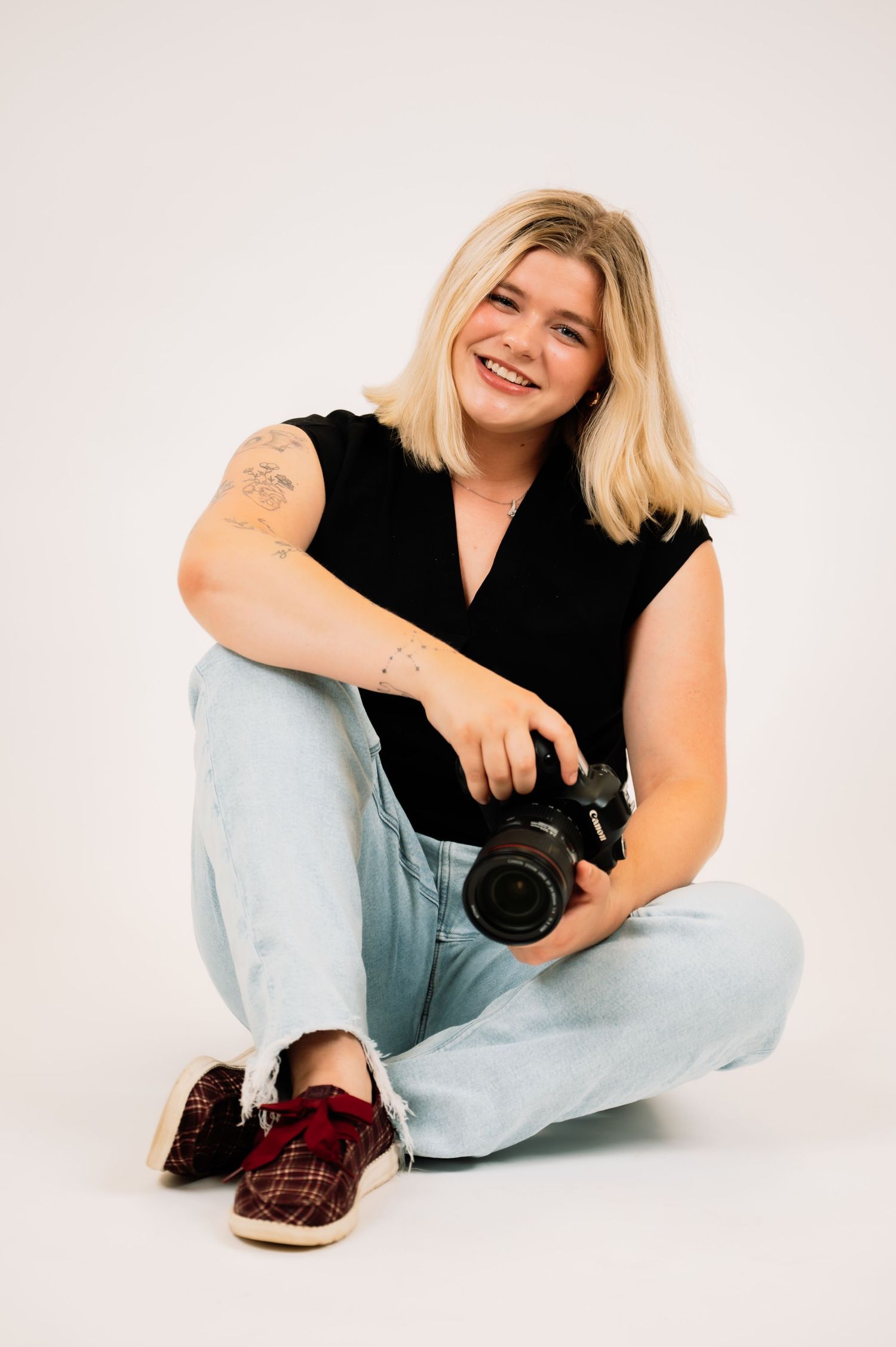Laughing tattooed blonde woman sitting cross-legged on floor holding DSLR camera in white studio.