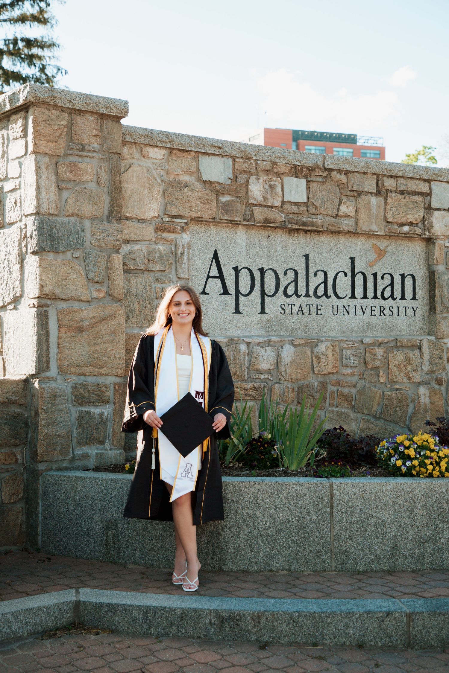 Female graduate in cap and gown standing before Appalachian State University stone sign.