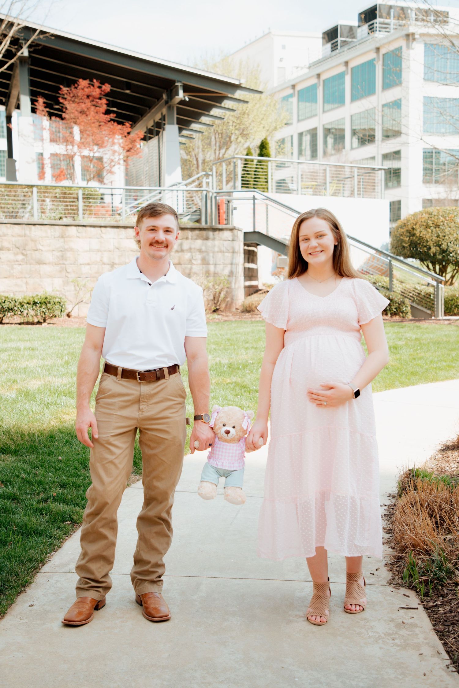 Expecting couple standing outdoors holding a teddy bear to announce baby girl pregnancy.