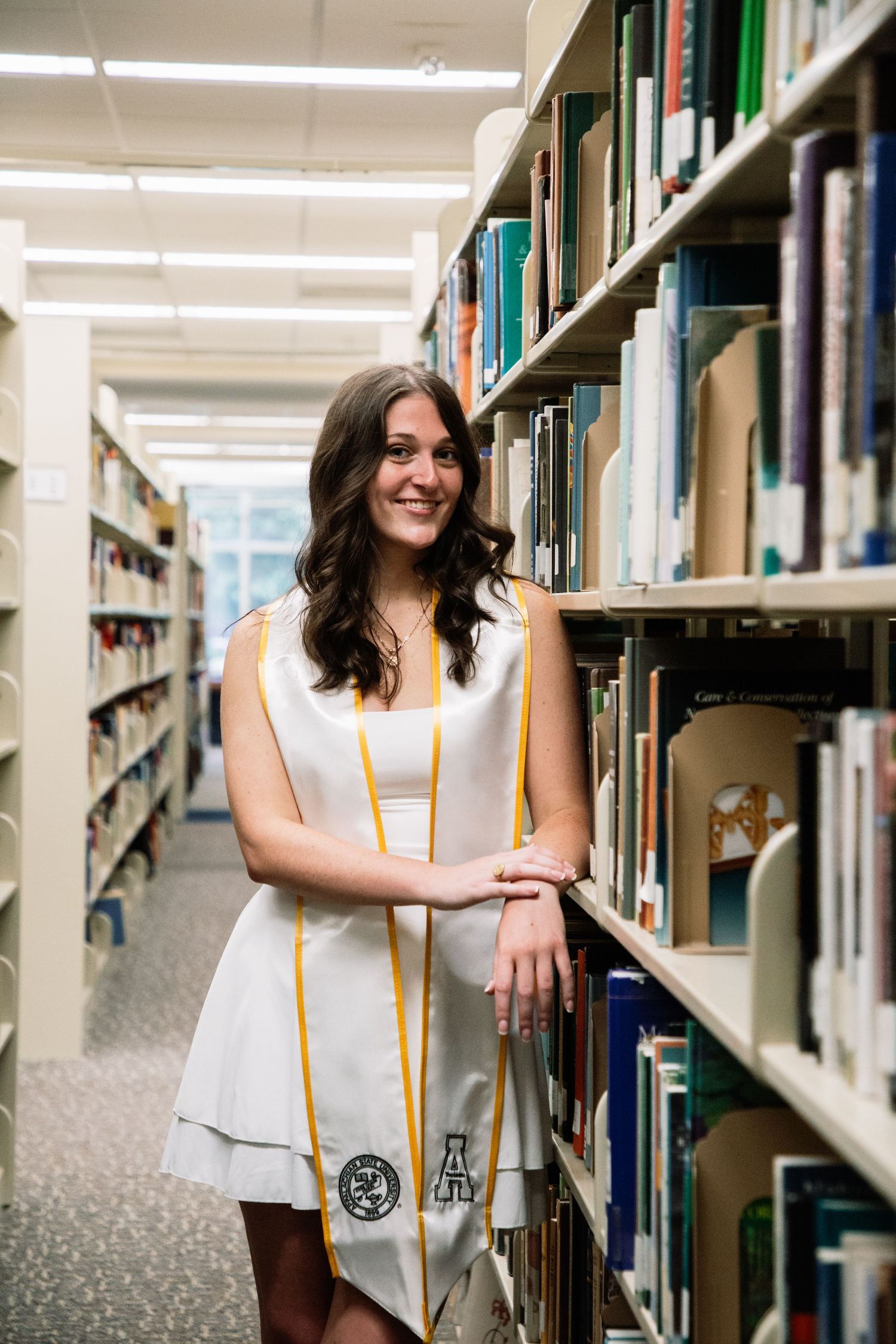 Female graduate in white dress and gold stole leans against library bookshelf smiling at camera.