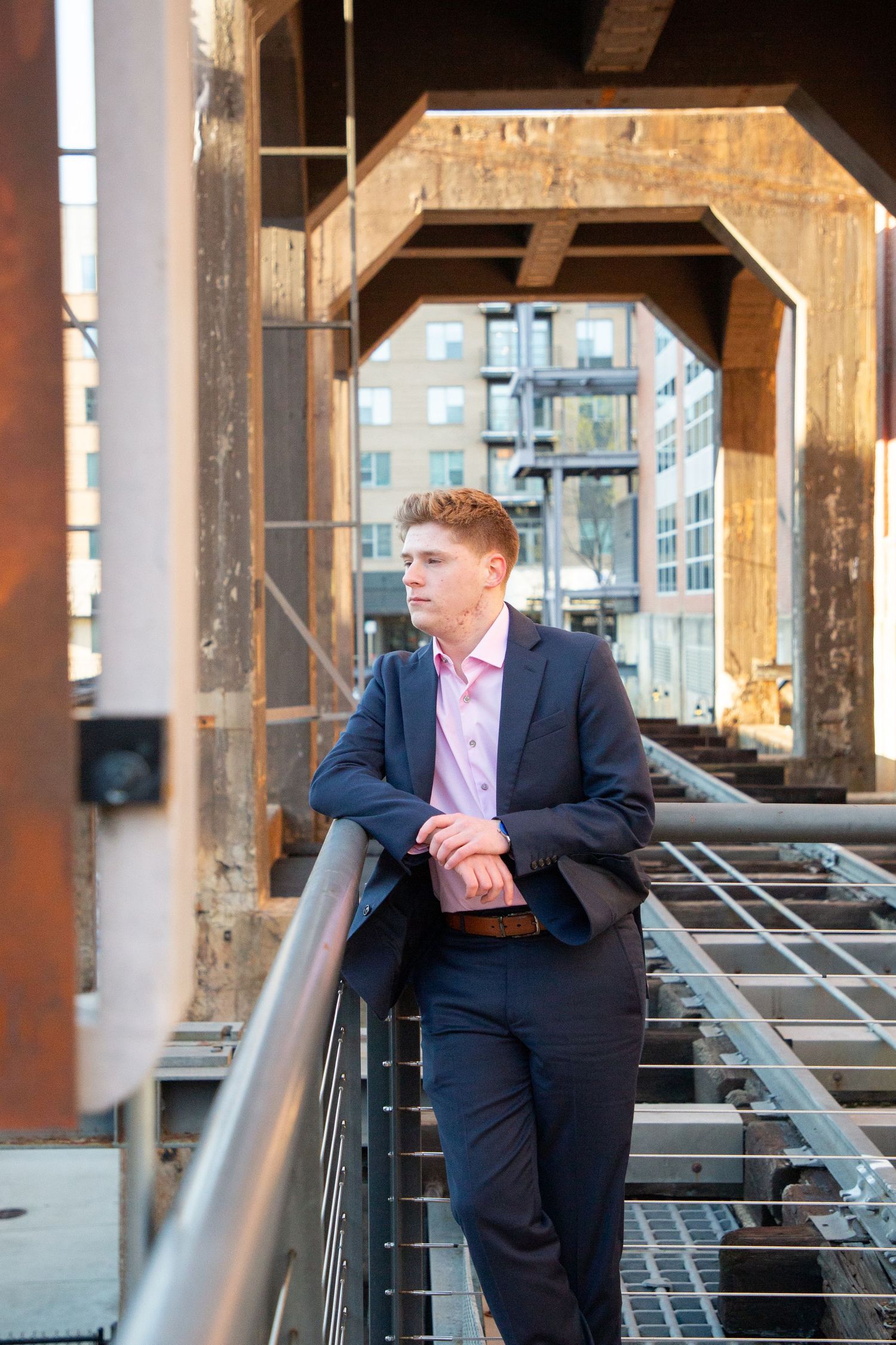 Young man in navy suit leaning on a railing inside a rustic industrial bridge structure.