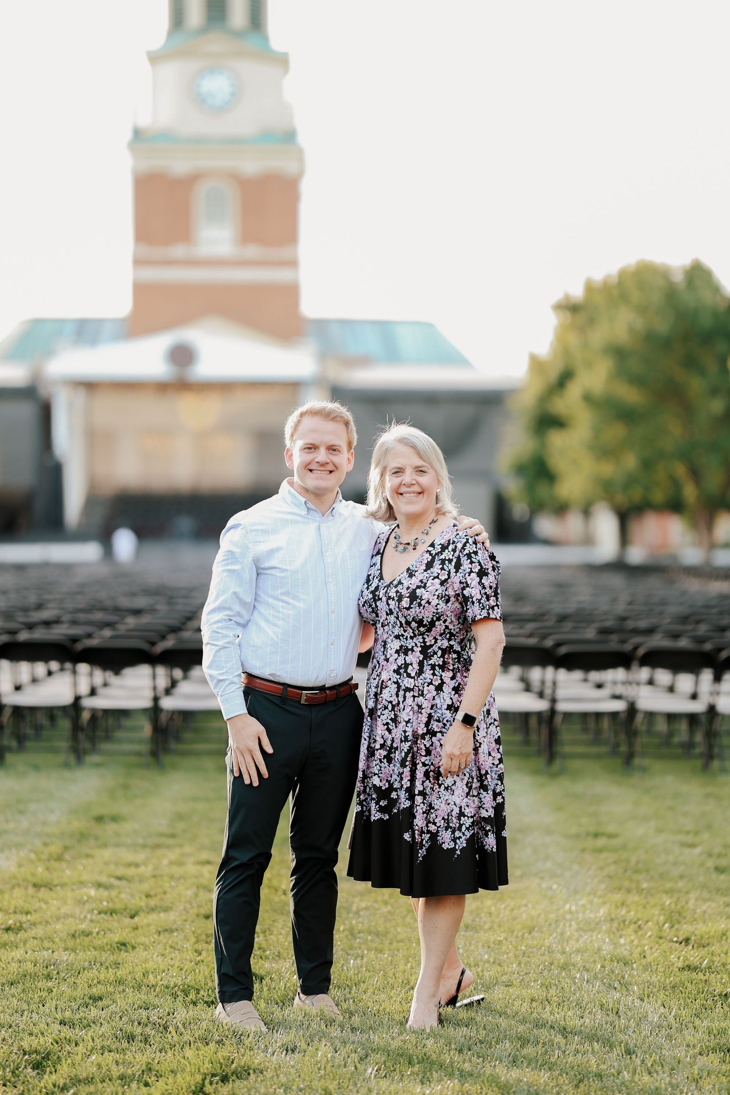 Young man and older woman smiling together outdoors with rows of black chairs and brick building behind.