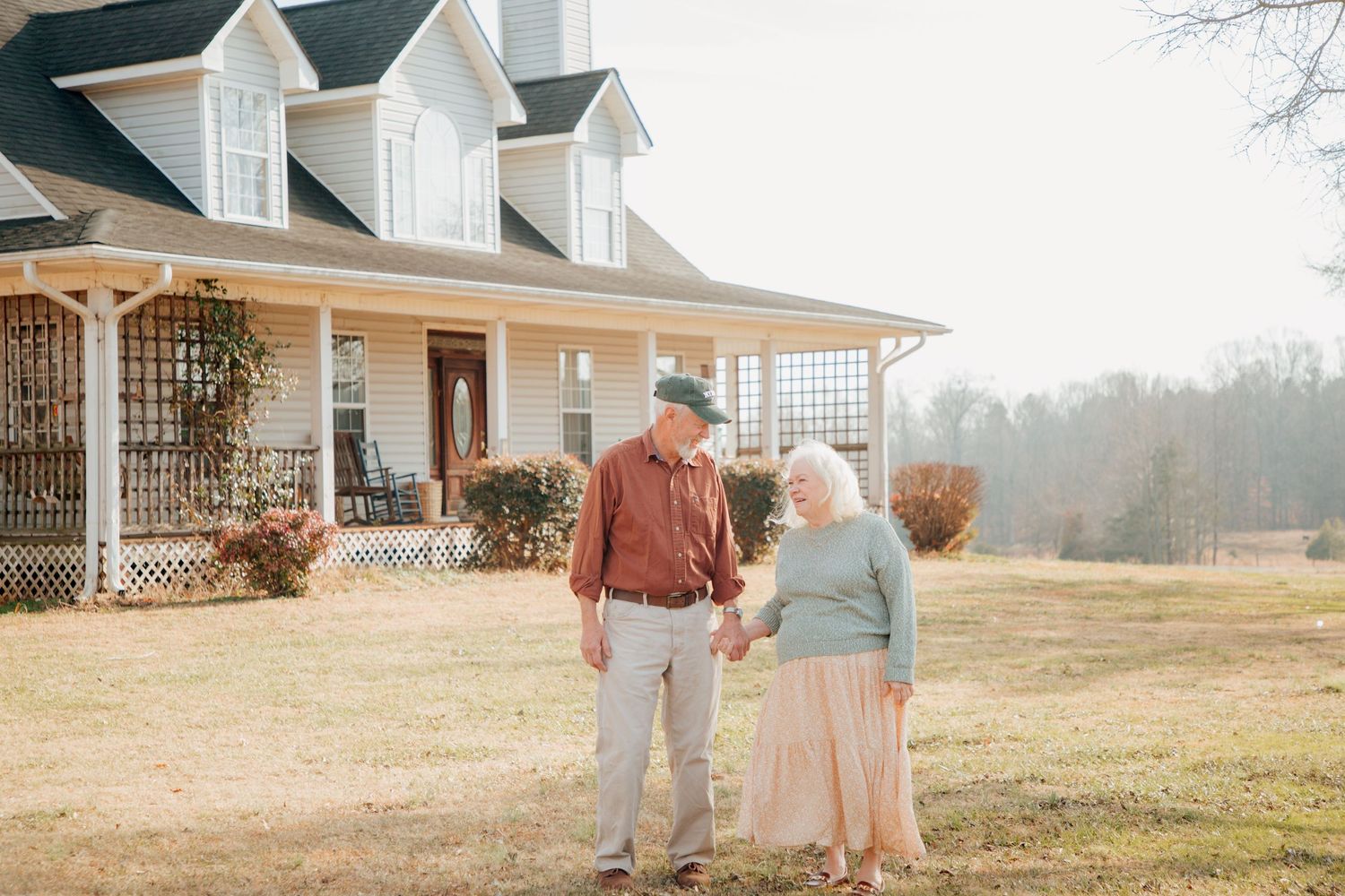 Loving elderly couple holding hands and gazing at each other before a white country home.