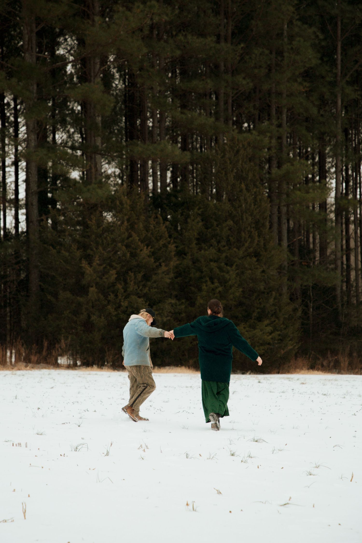 Couple running playfully hand in hand across snowy field toward dense pine forest in winter.