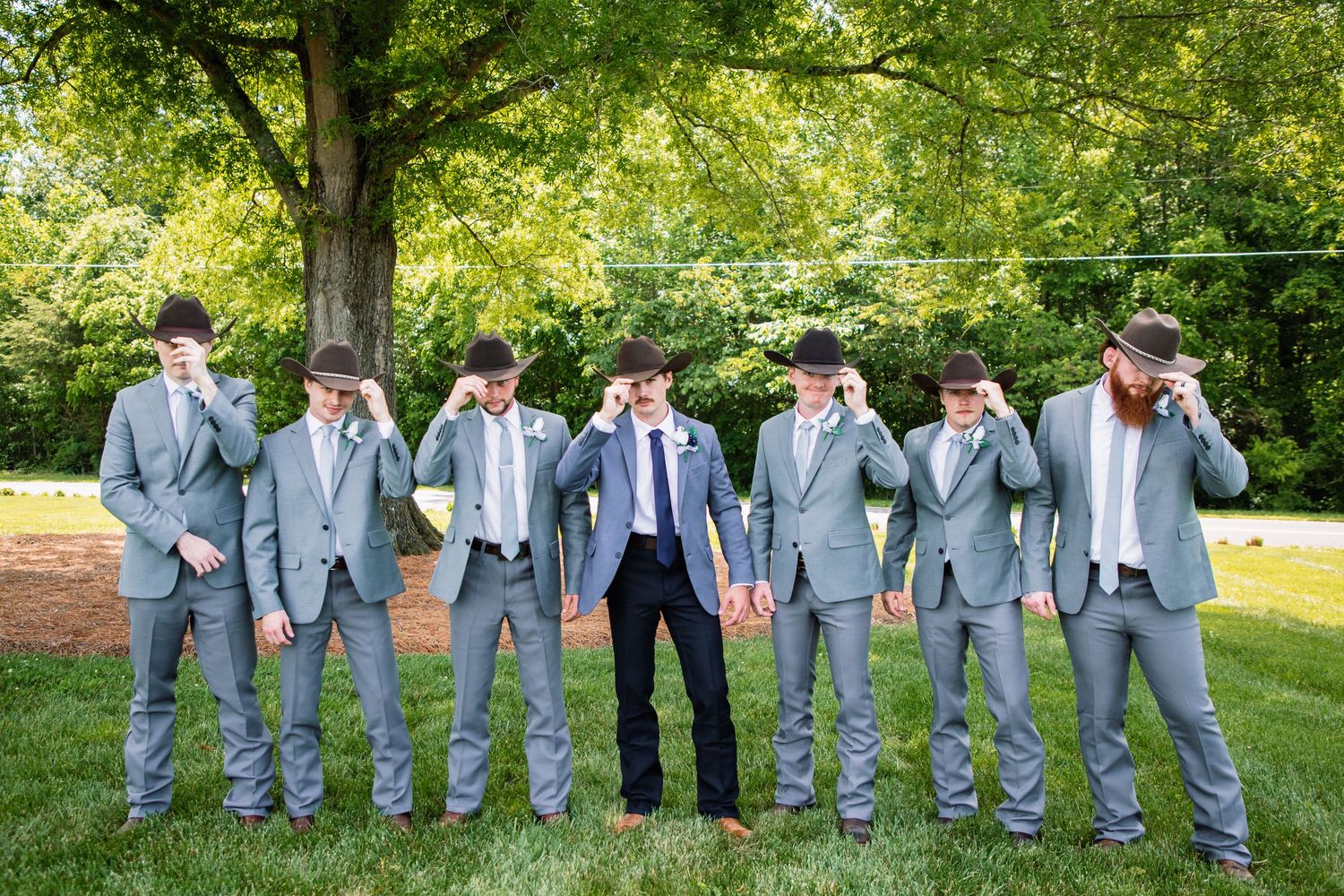 Groom and groomsmen in gray suits and cowboy hats tip their hats in a fun coordinated outdoor wedding pose.