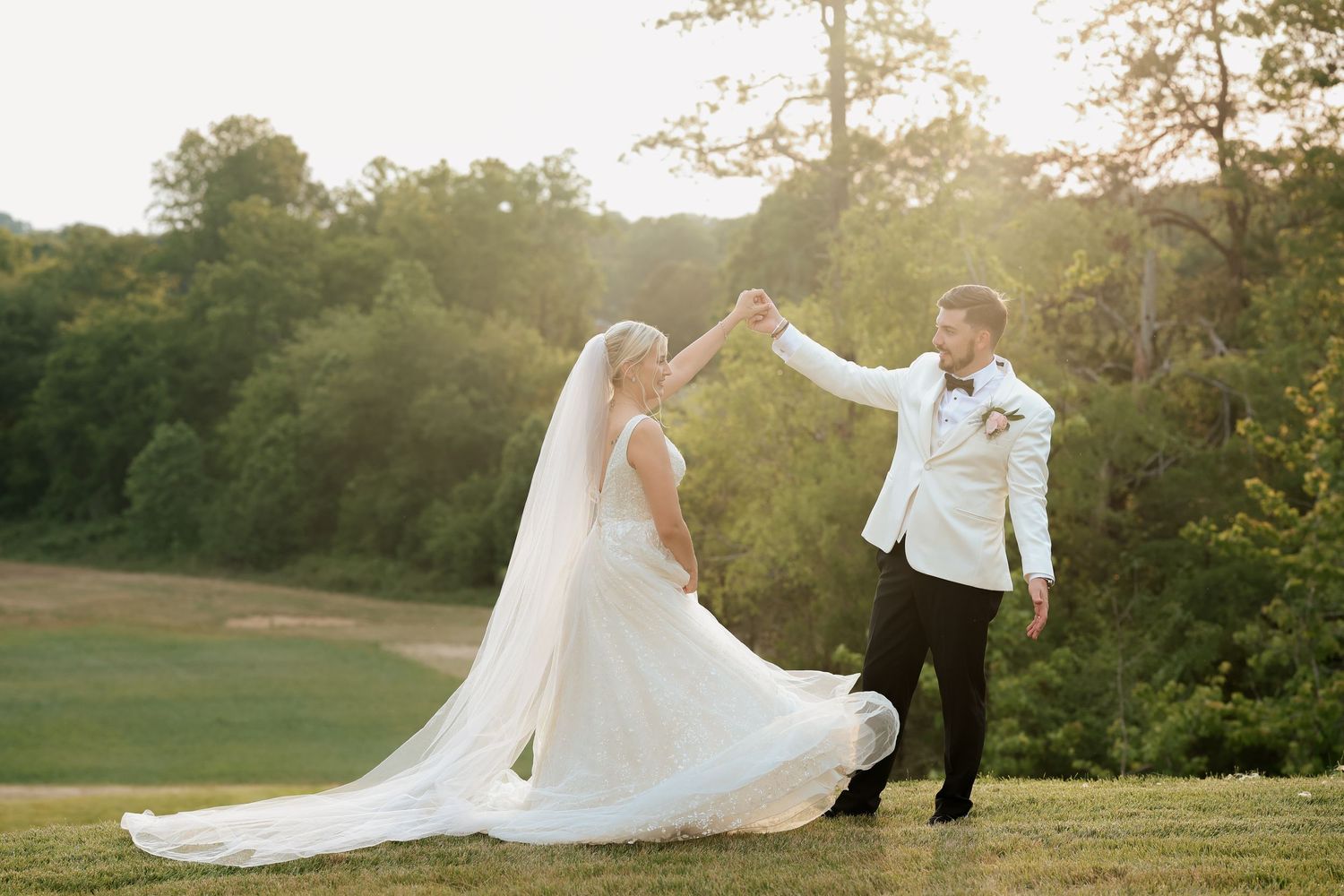 Groom twirling bride in flowing white wedding gown and veil on a sunlit green lawn at sunset.