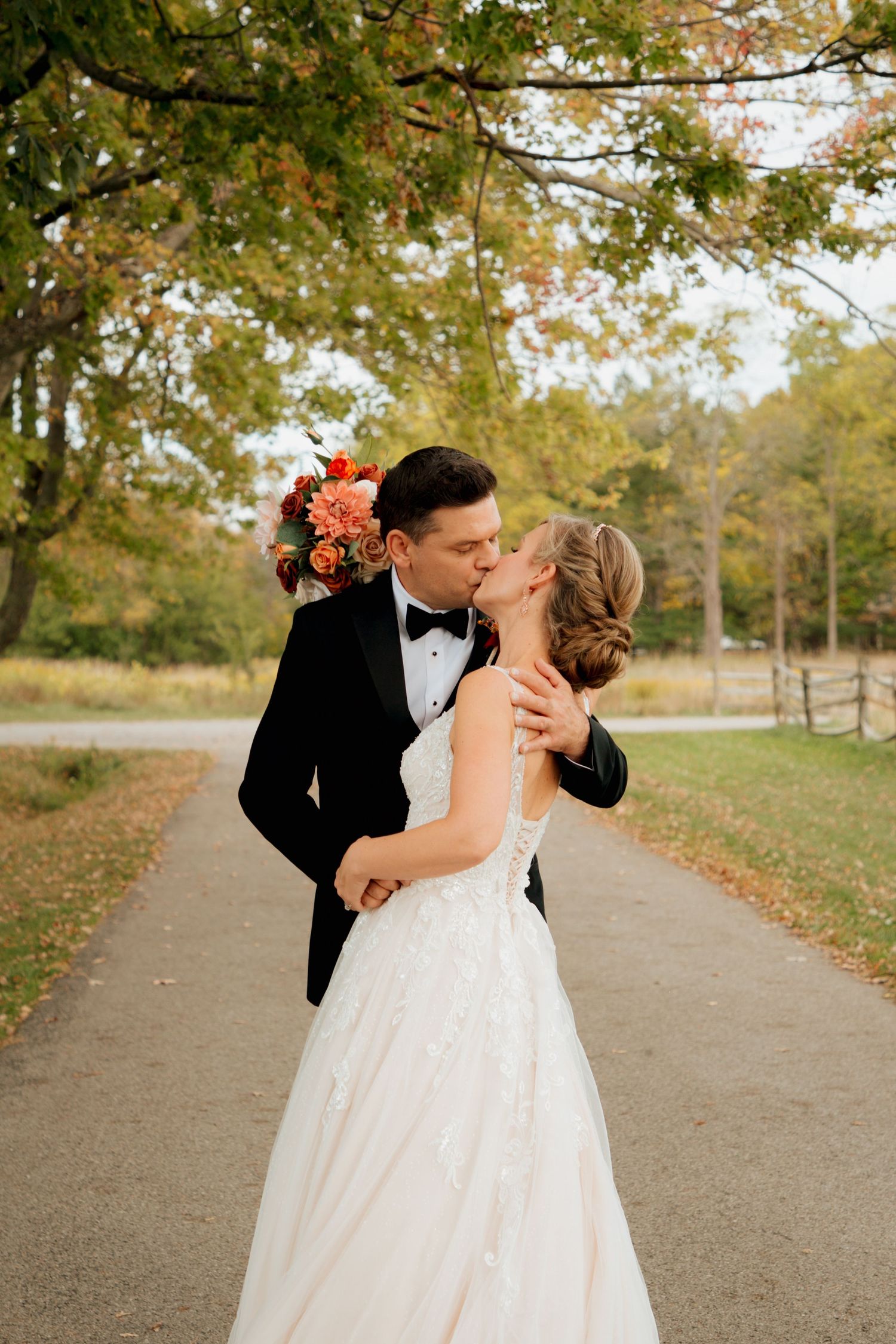 Bride and groom share romantic kiss on tree-lined path during autumn wedding portraits.
