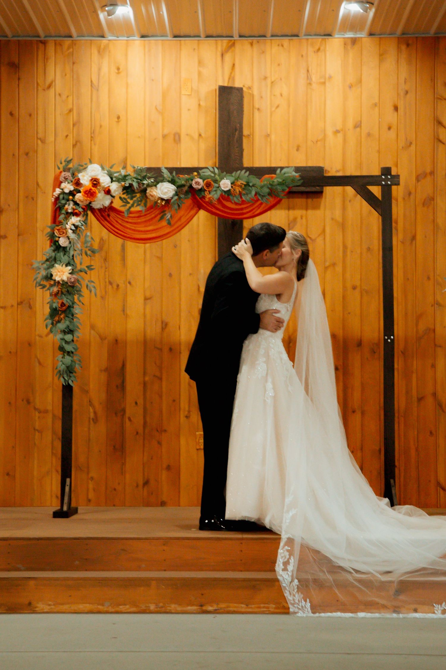 Bride and groom share first kiss beneath floral-adorned wooden cross arch at rustic chapel.