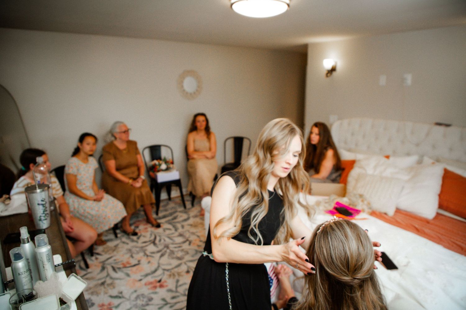 Hair stylist in black dress styling bridesmaid's hair while bridal party watches in decorated room.