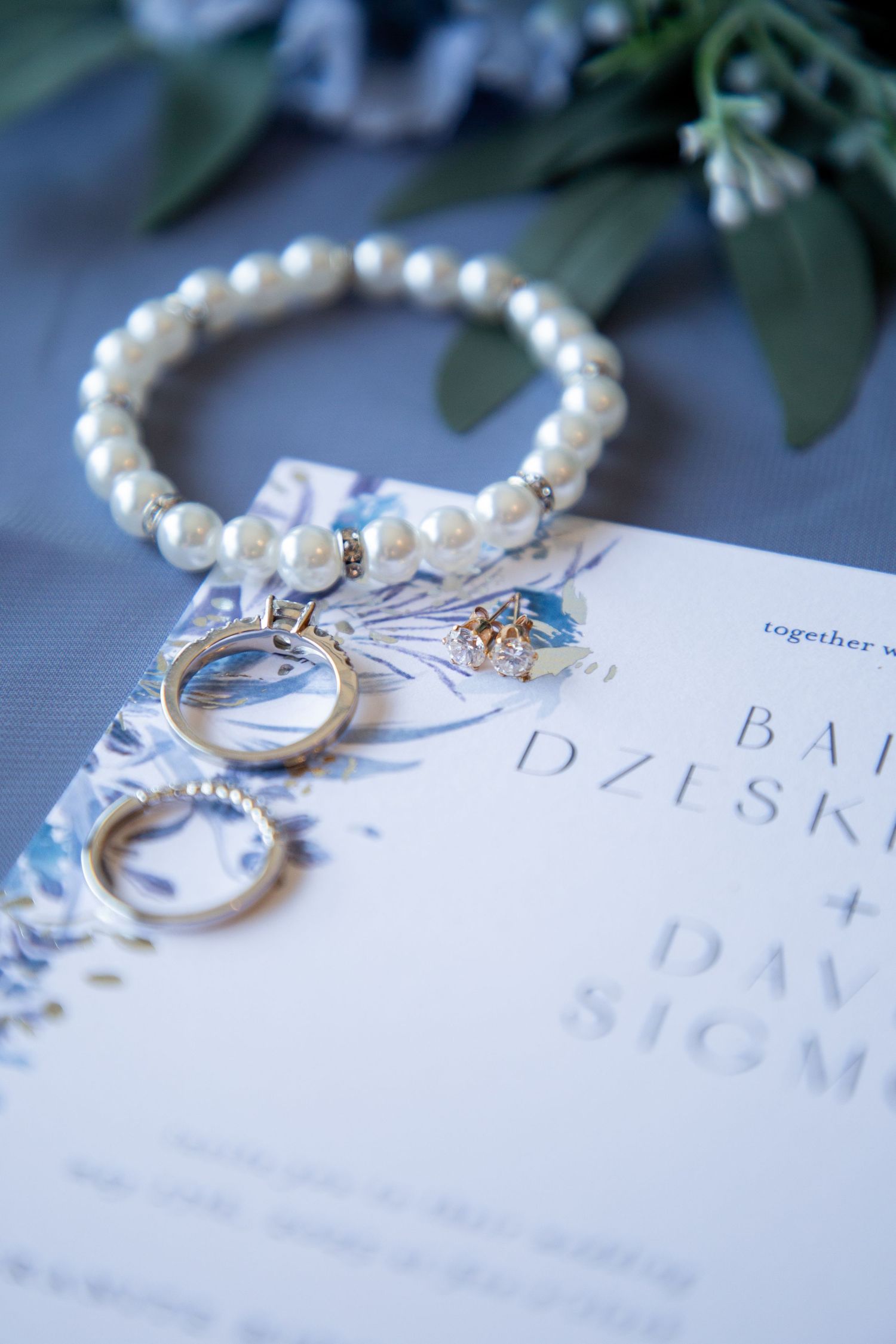 Pearl bracelet and gold jewelry pieces arranged on a blue floral wedding invitation on gray fabric.