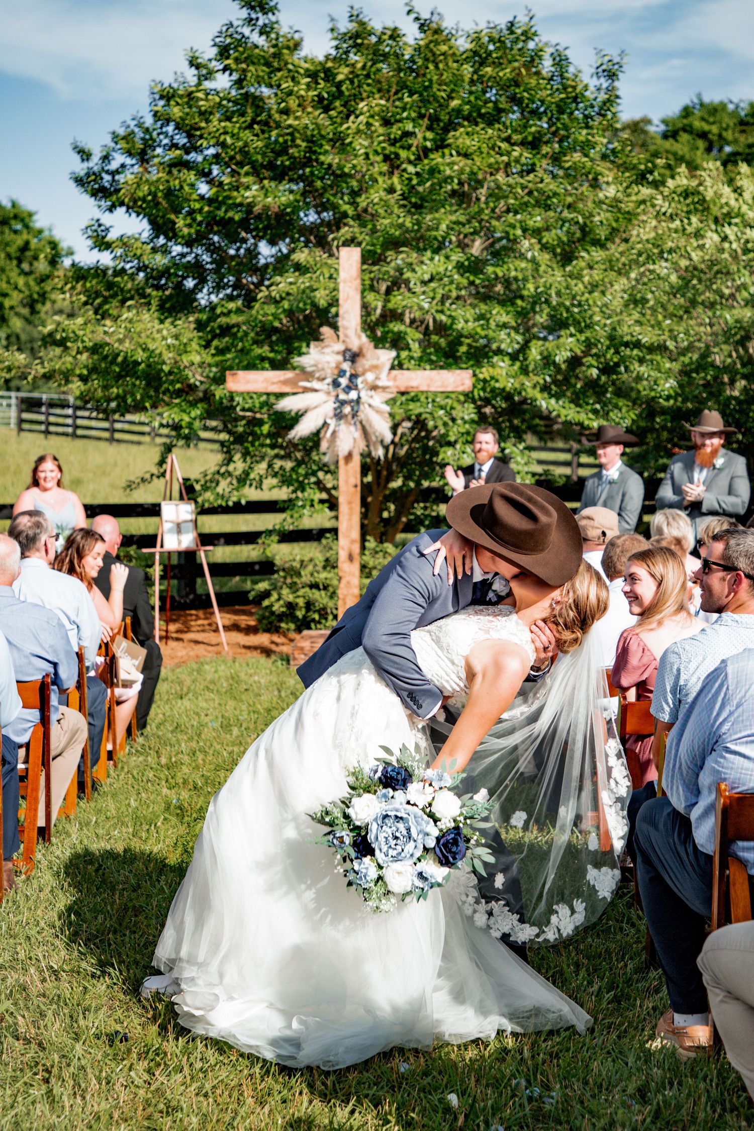 Groom in cowboy hat dips bride in lace gown for a kiss while walking down outdoor wedding aisle.