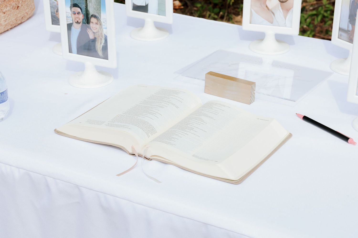 Open guest book with pencil and framed photos on white linen table at outdoor wedding reception.