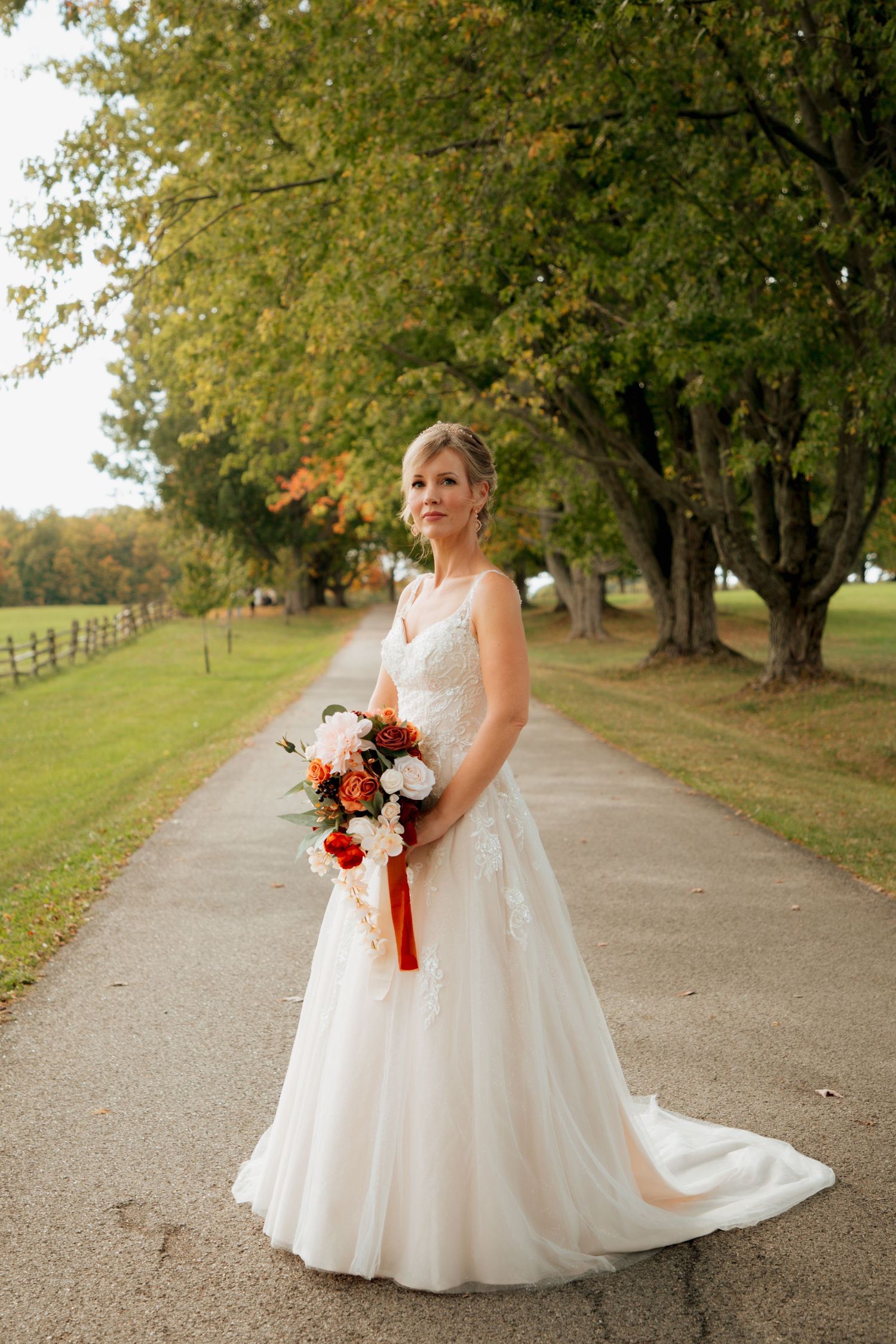 Bride in a flowing lace wedding dress stands on a scenic tree-lined path holding an autumn floral bouquet.