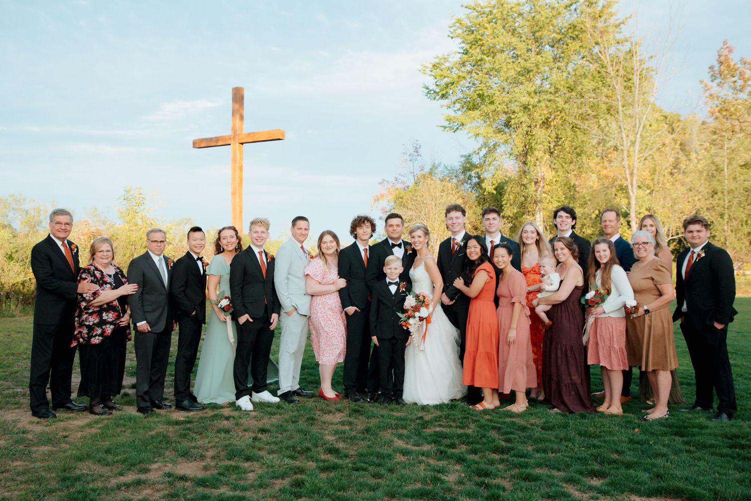 Large wedding party and family group portrait outdoors beside a tall wooden cross on a sunny autumn day.