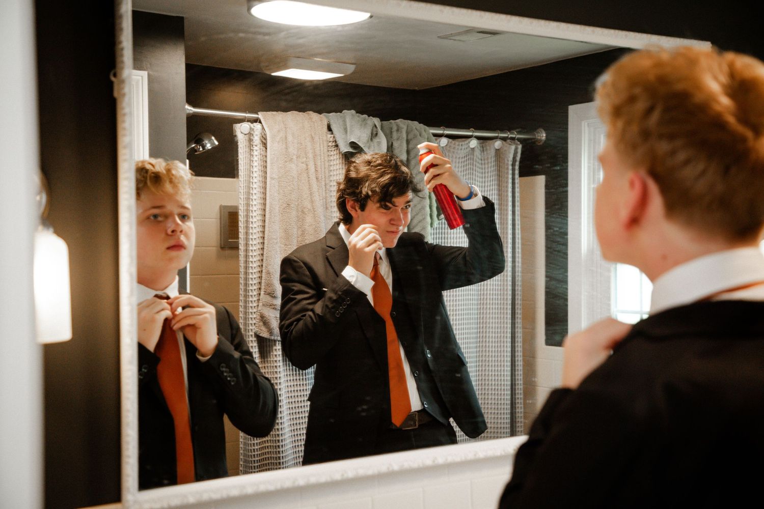 Young man in suit adjusting orange tie while looking in a dark-framed mirror.