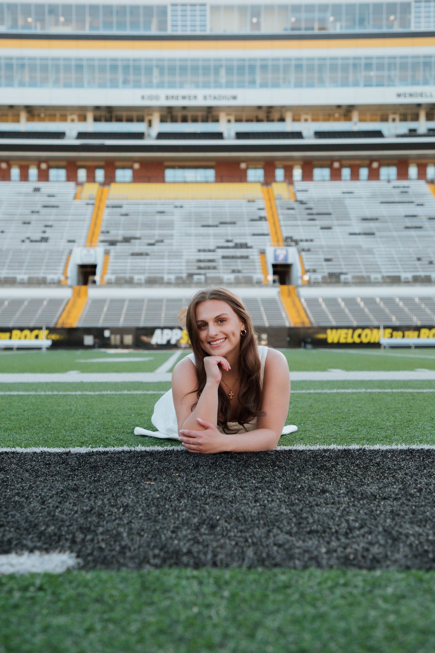 Smiling graduate in white dress poses on turf at Kidd Brewer Stadium football field.