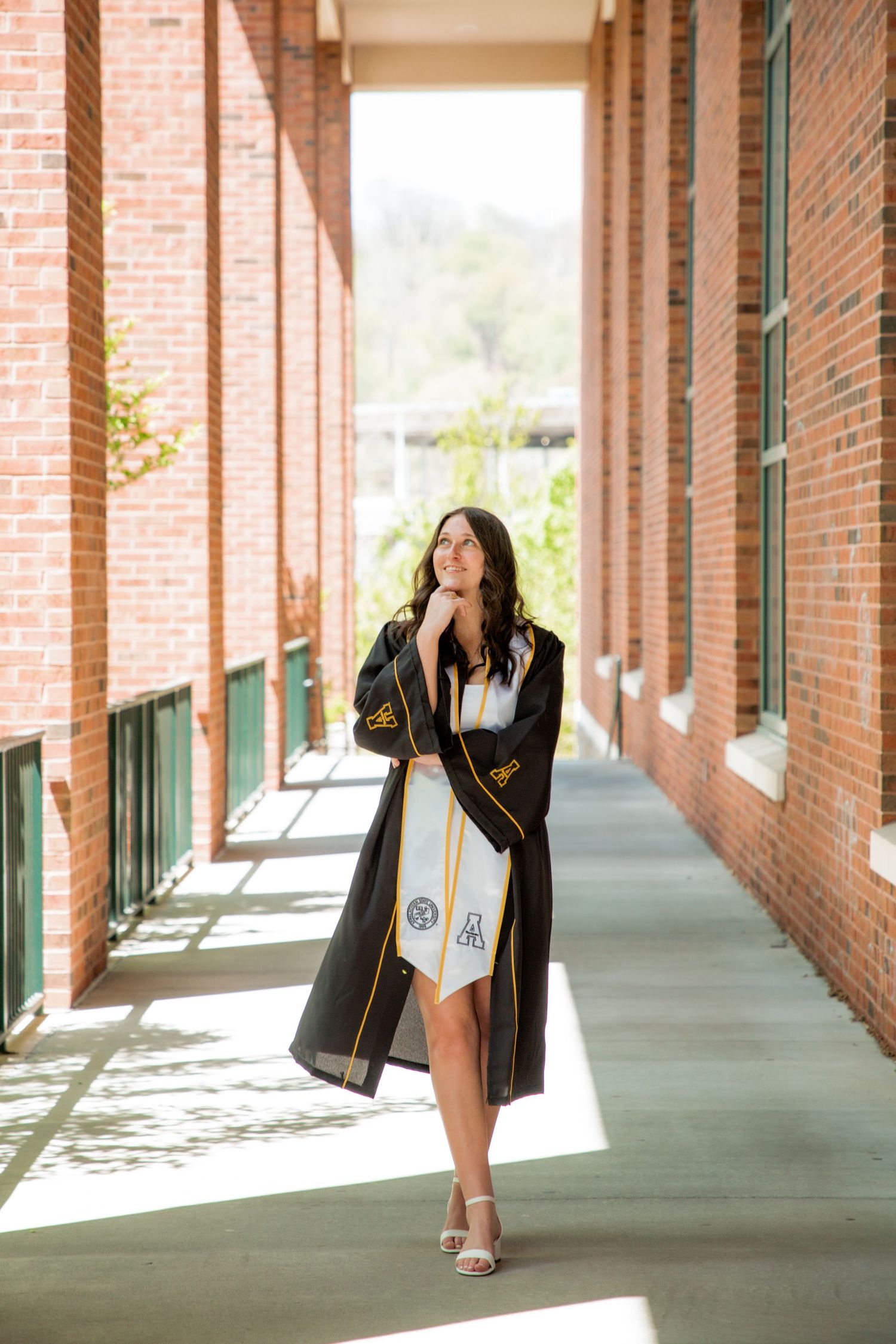 Female graduate in black robe posing thoughtfully in sunny brick hallway.