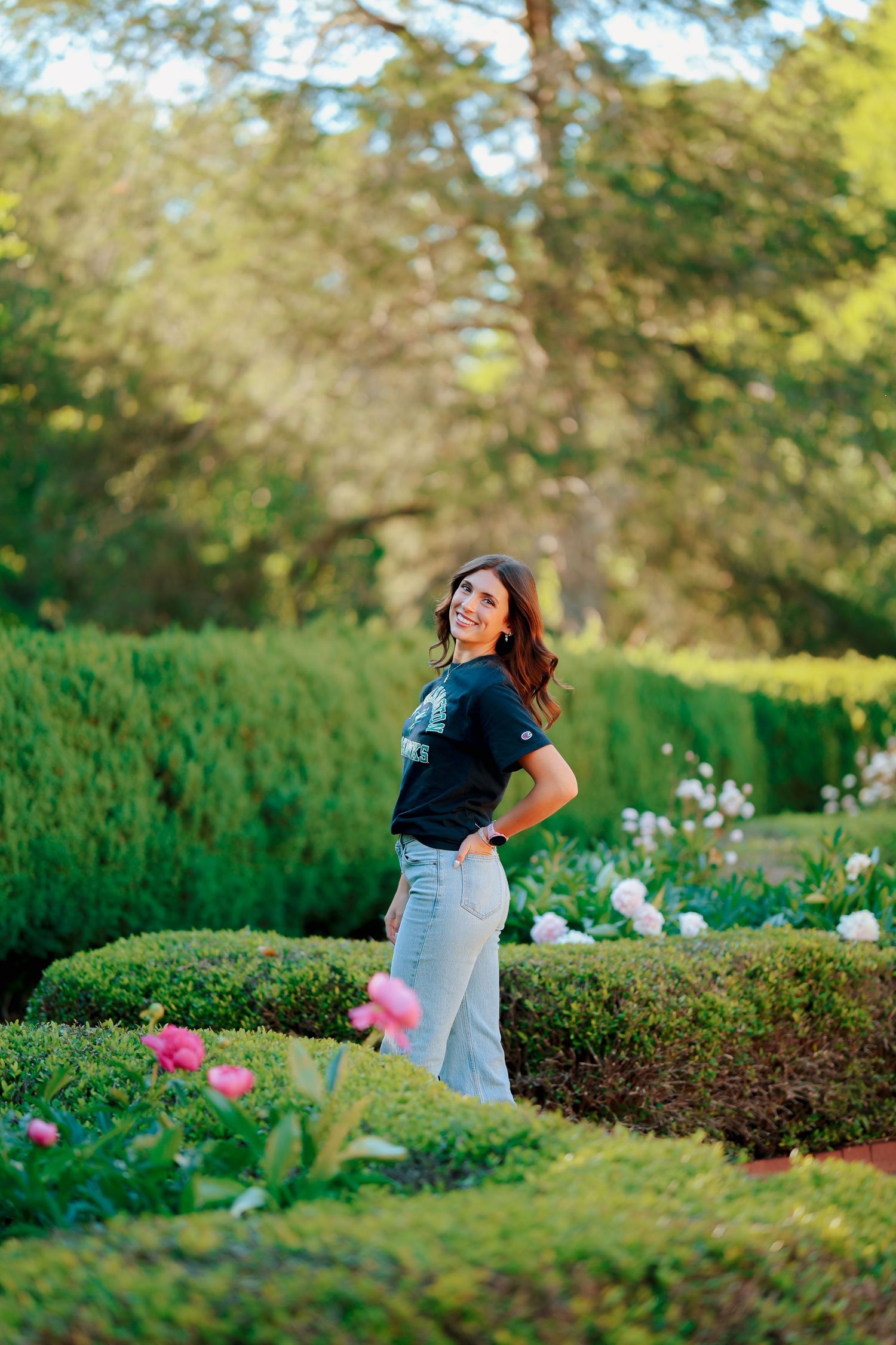 Young woman in UNCW Seahawks shirt smiling over shoulder while standing in a lush garden with pink flowers.