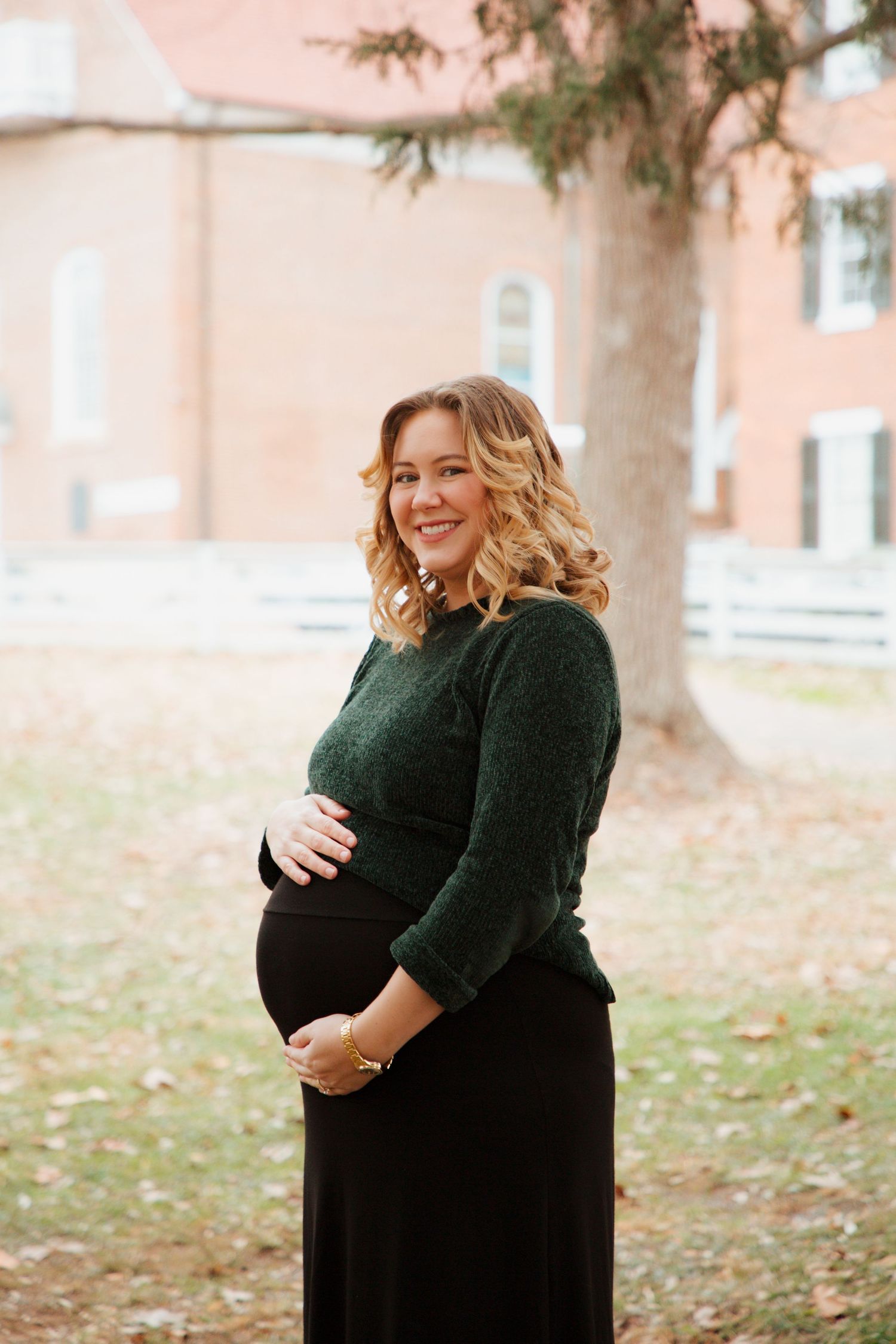 Smiling pregnant woman in green sweater and black skirt cradling baby bump outdoors.
