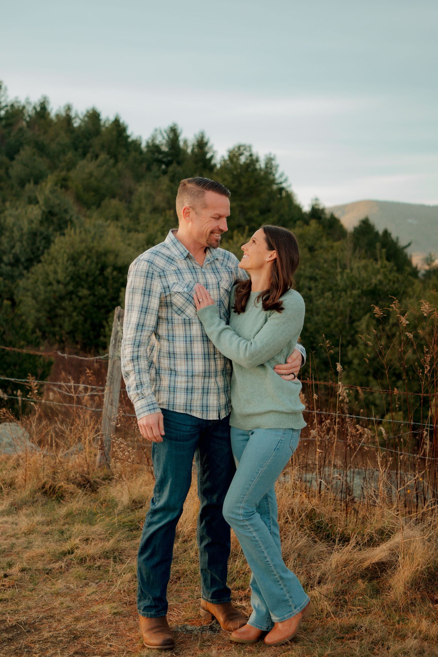 Couple gazes at each other lovingly in golden hour light with green forest and mountain backdrop.