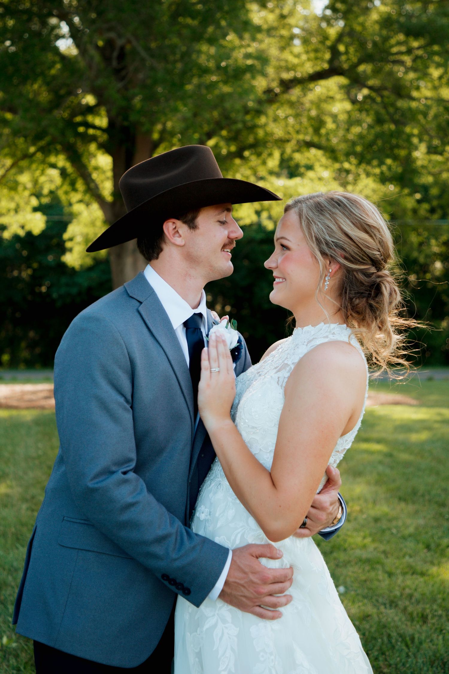 Smiling bride in lace gown embracing groom in gray suit and black cowboy hat outdoors.