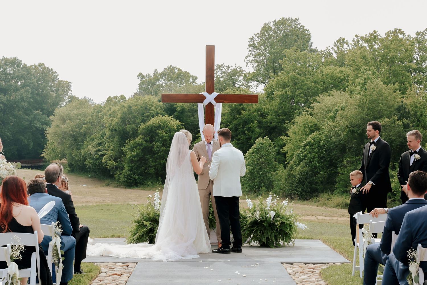 Outdoor wedding ceremony with couple at altar before large wooden cross, guests seated in white chairs on lush green grounds.