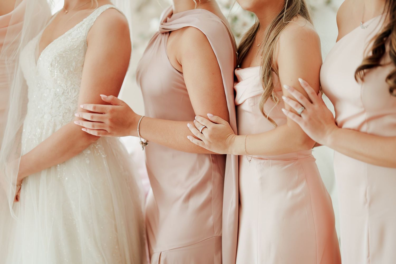 Bride and bridesmaids in blush dresses posing closely together showing elegant jewelry details.