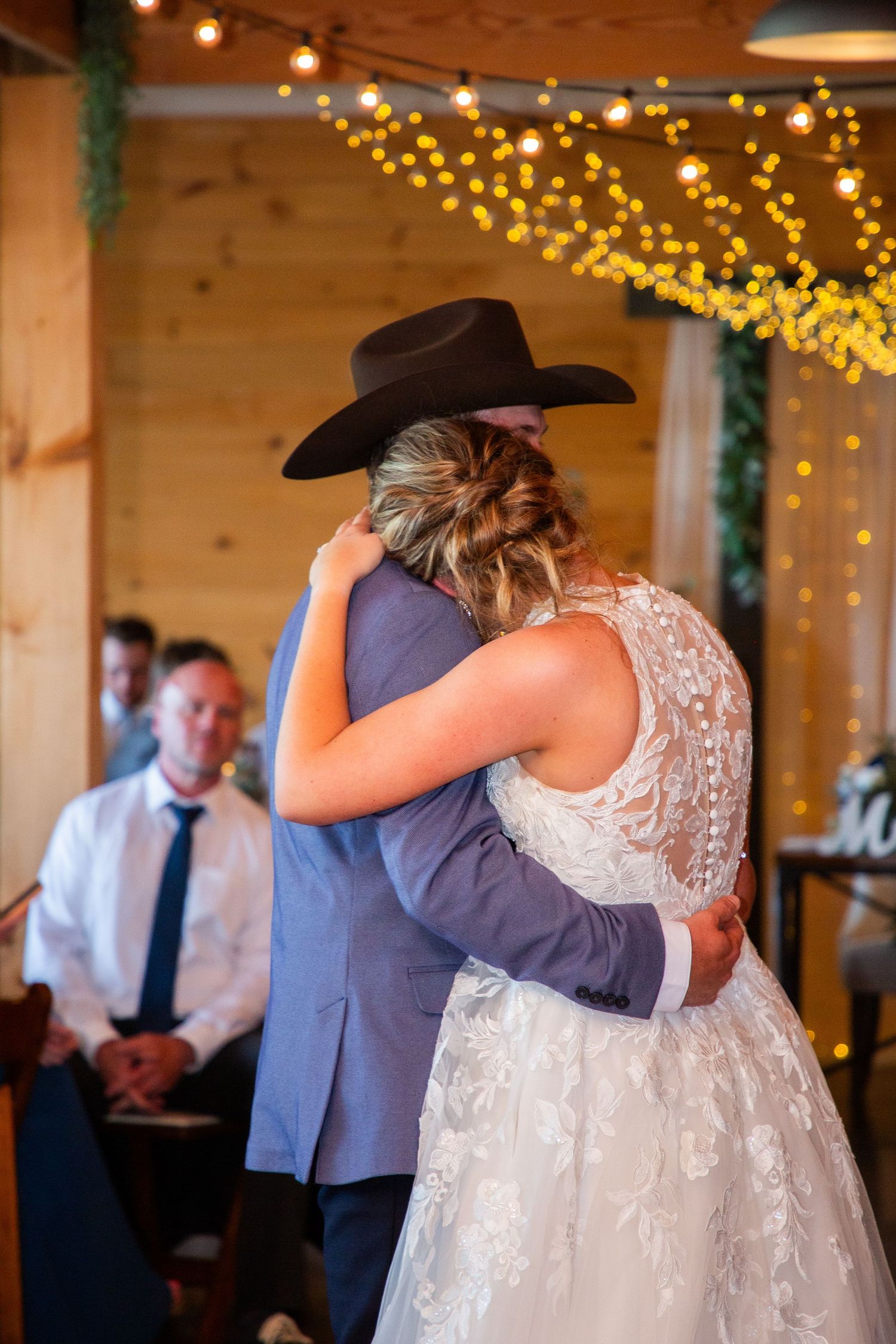 Newlyweds embrace during their first dance in a warmly lit barn reception venue with bokeh string lights.