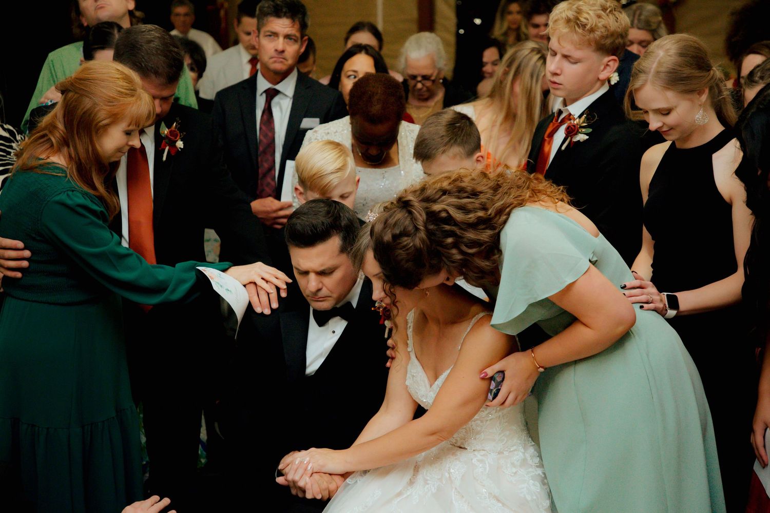 Wedding party and guests surround kneeling bride and groom in a circle of prayer.