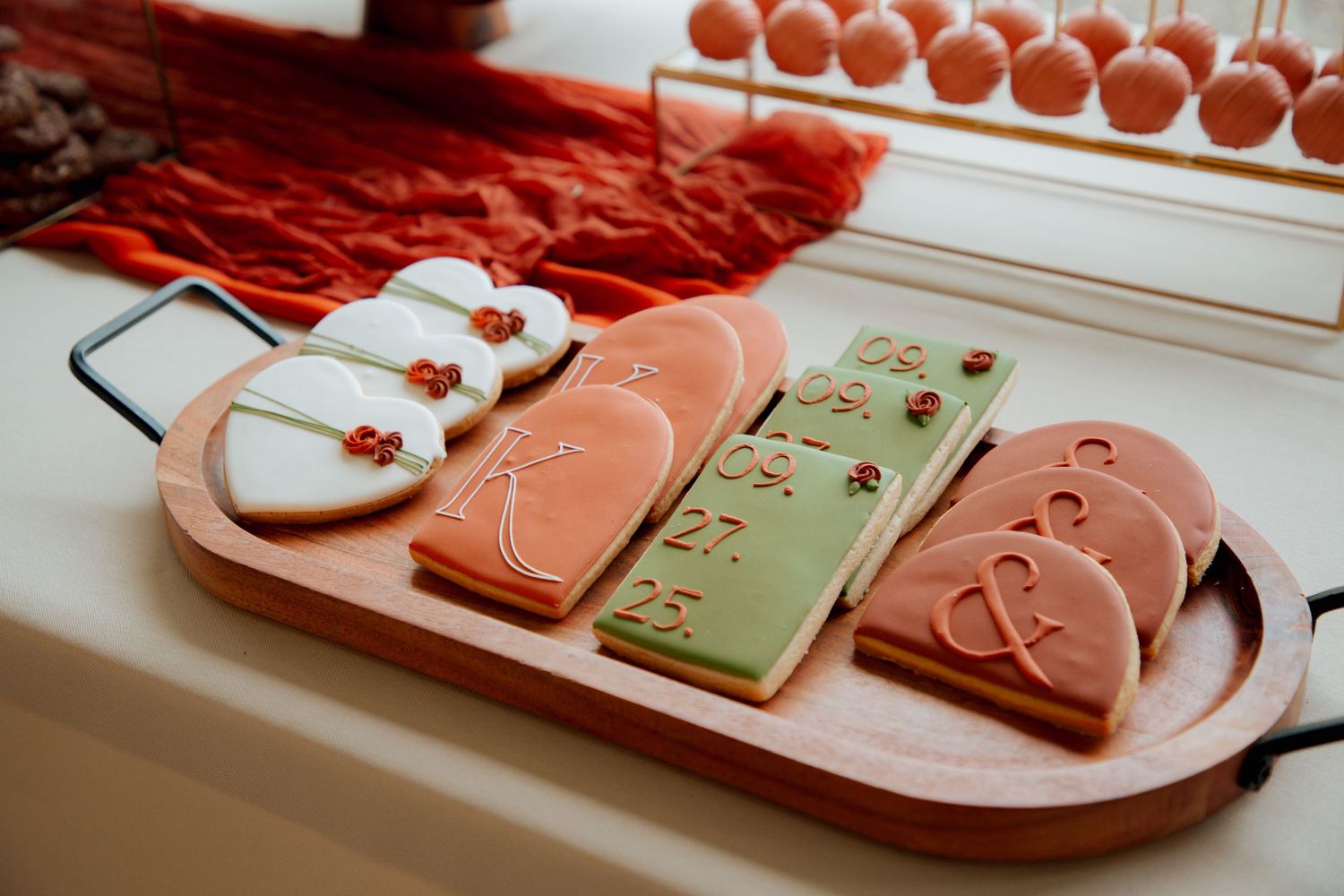 Decorated wedding sugar cookies on wooden board featuring hearts, ampersand, and date in pink, white, and green icing.