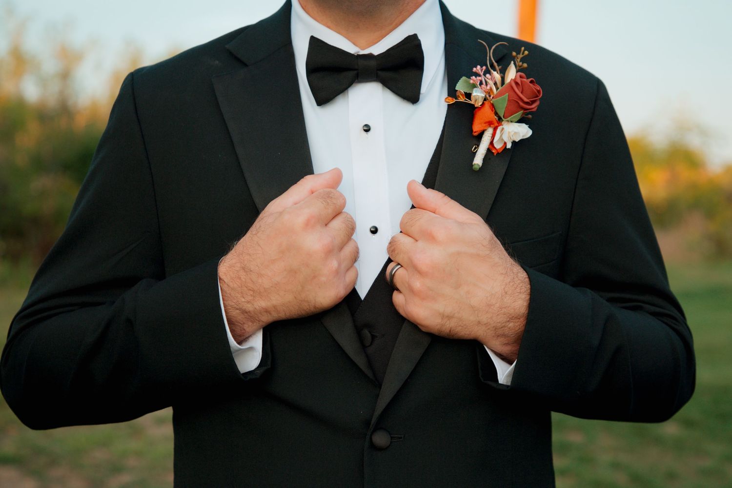 Close-up of groom adjusting black tuxedo with colorful floral boutonniere and bow tie.