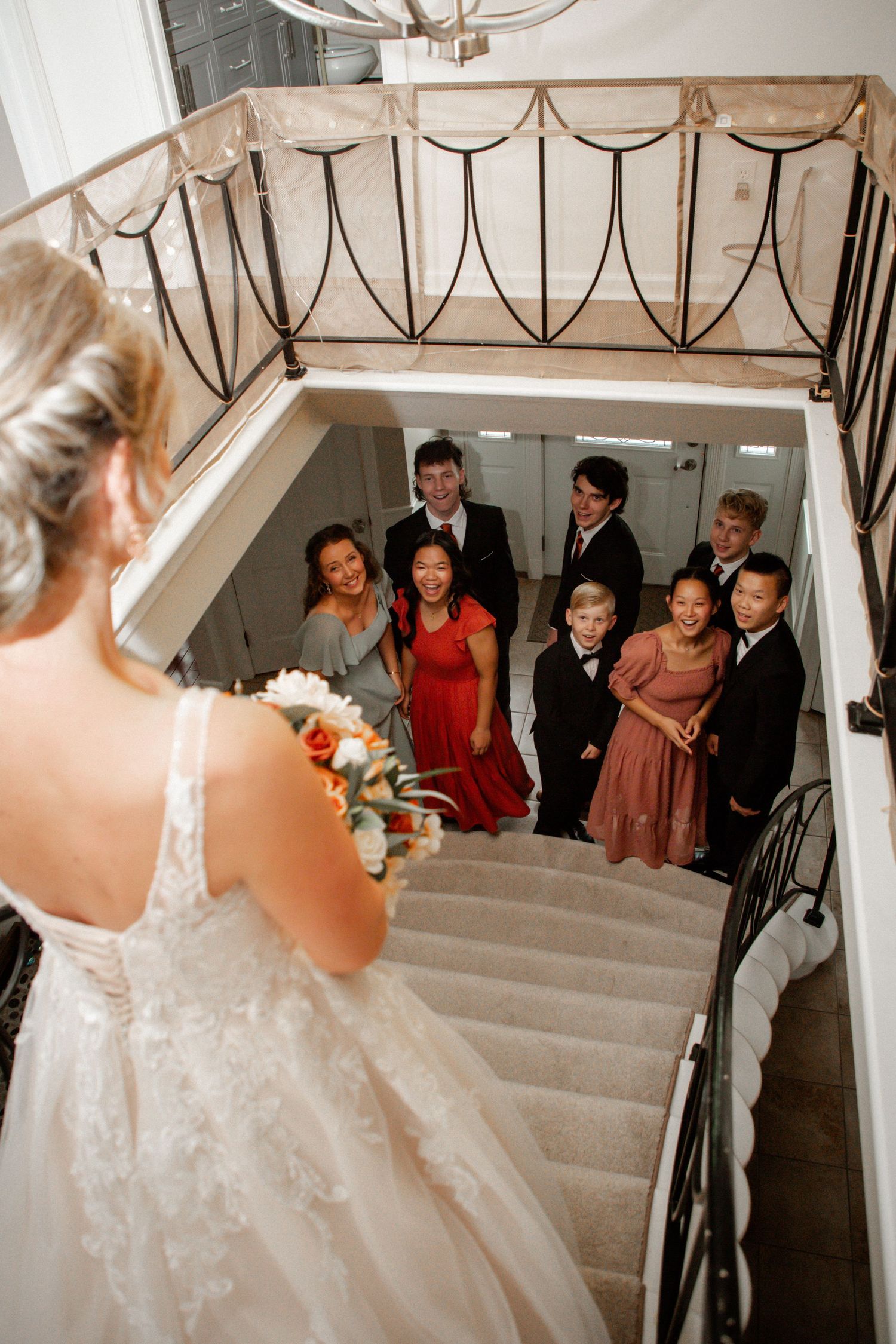 Bride in white lace gown holds bouquet atop elegant staircase as wedding party looks up in admiration.