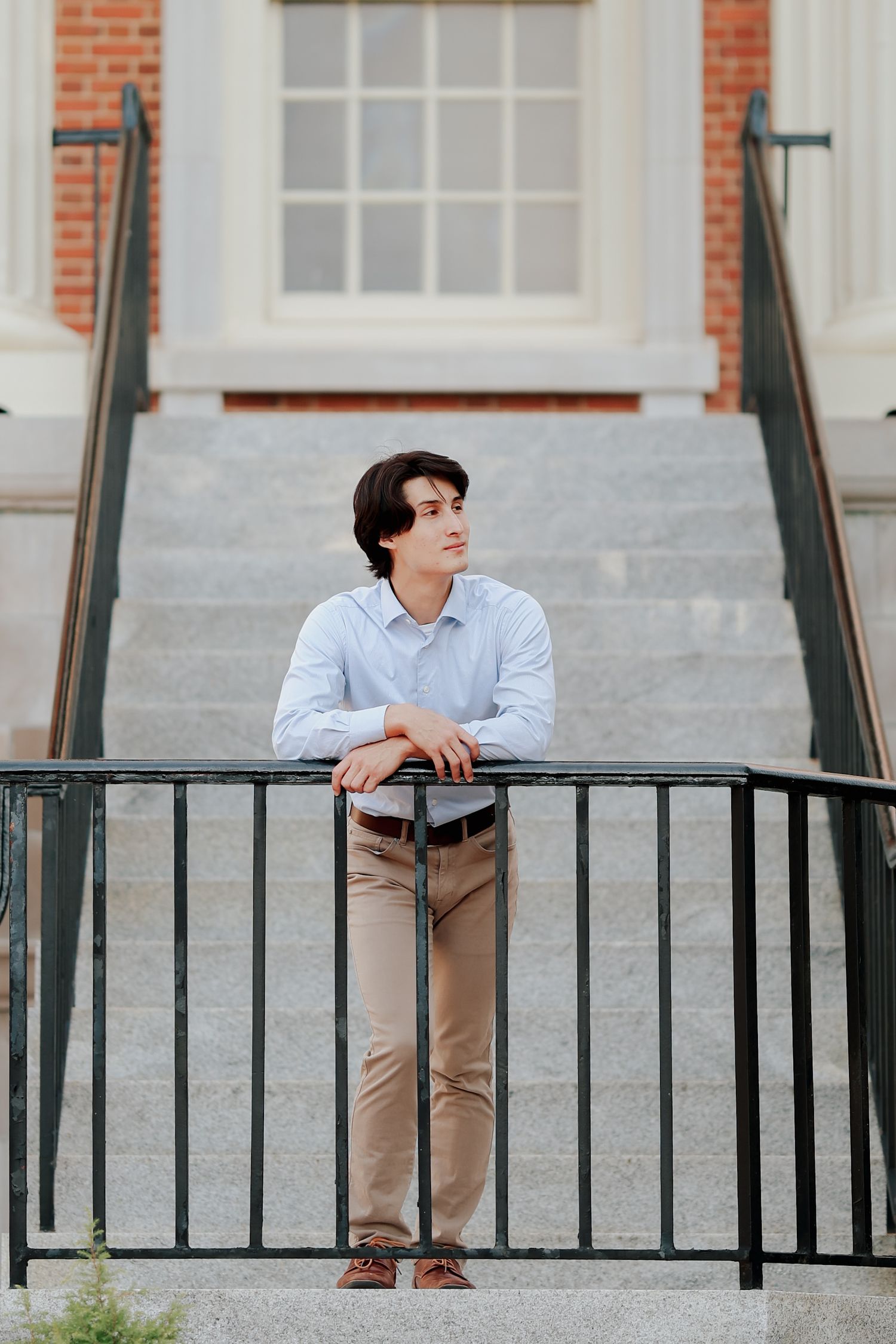 Young man in blue shirt leans on black iron railing on stone steps outside a brick building.