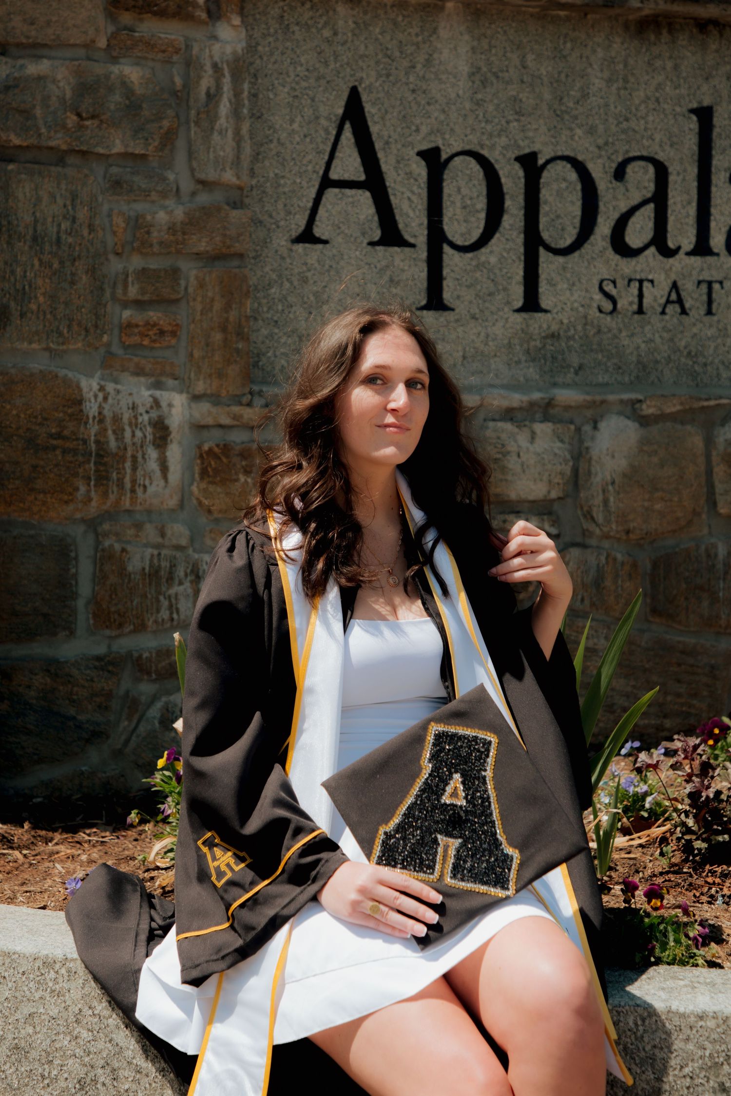 Female graduate holding decorated cap with rhinestone A seated at Appalachian State University sign.