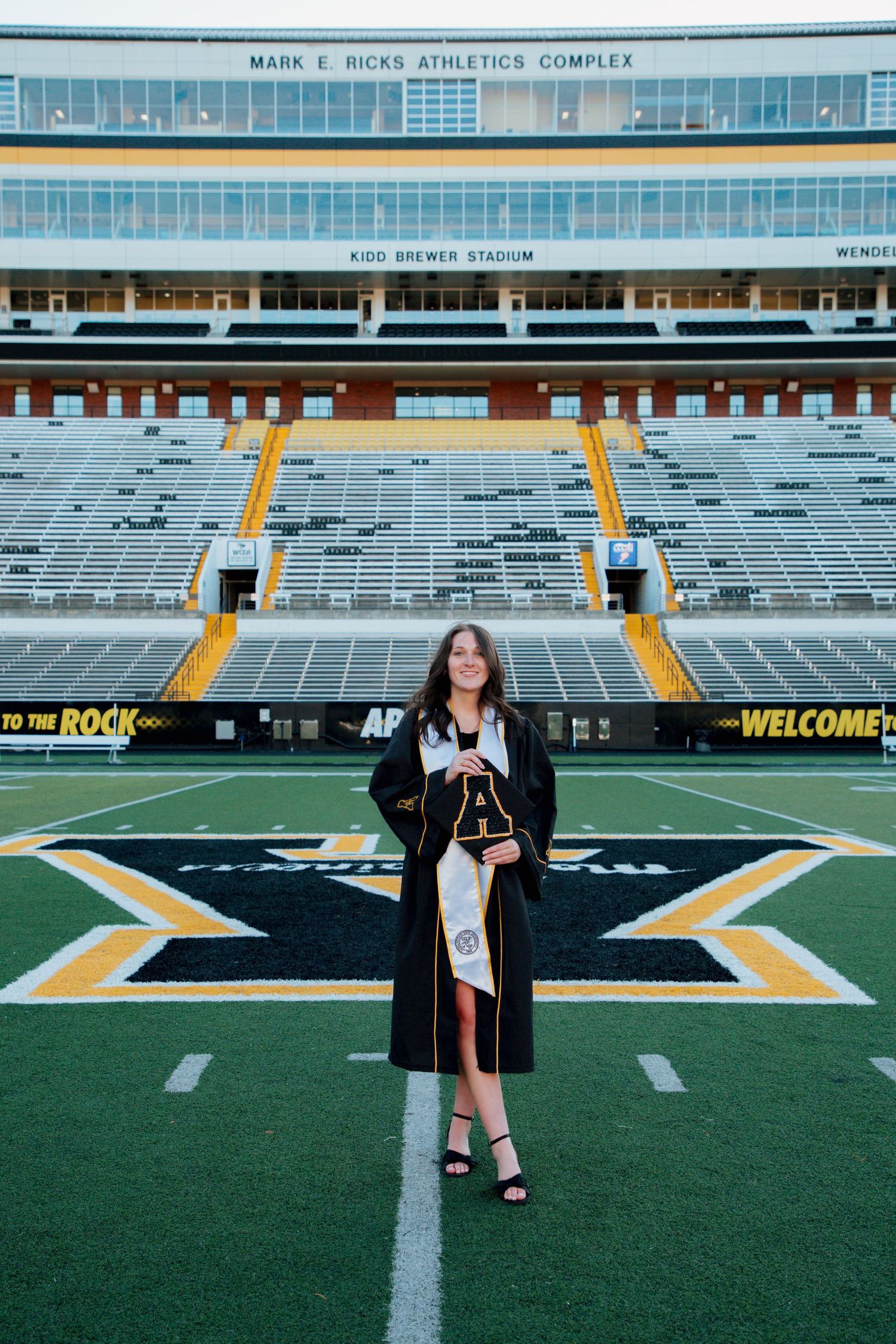 Graduate in black cap and gown standing on App State Kidd Brewer Stadium field holding diploma.