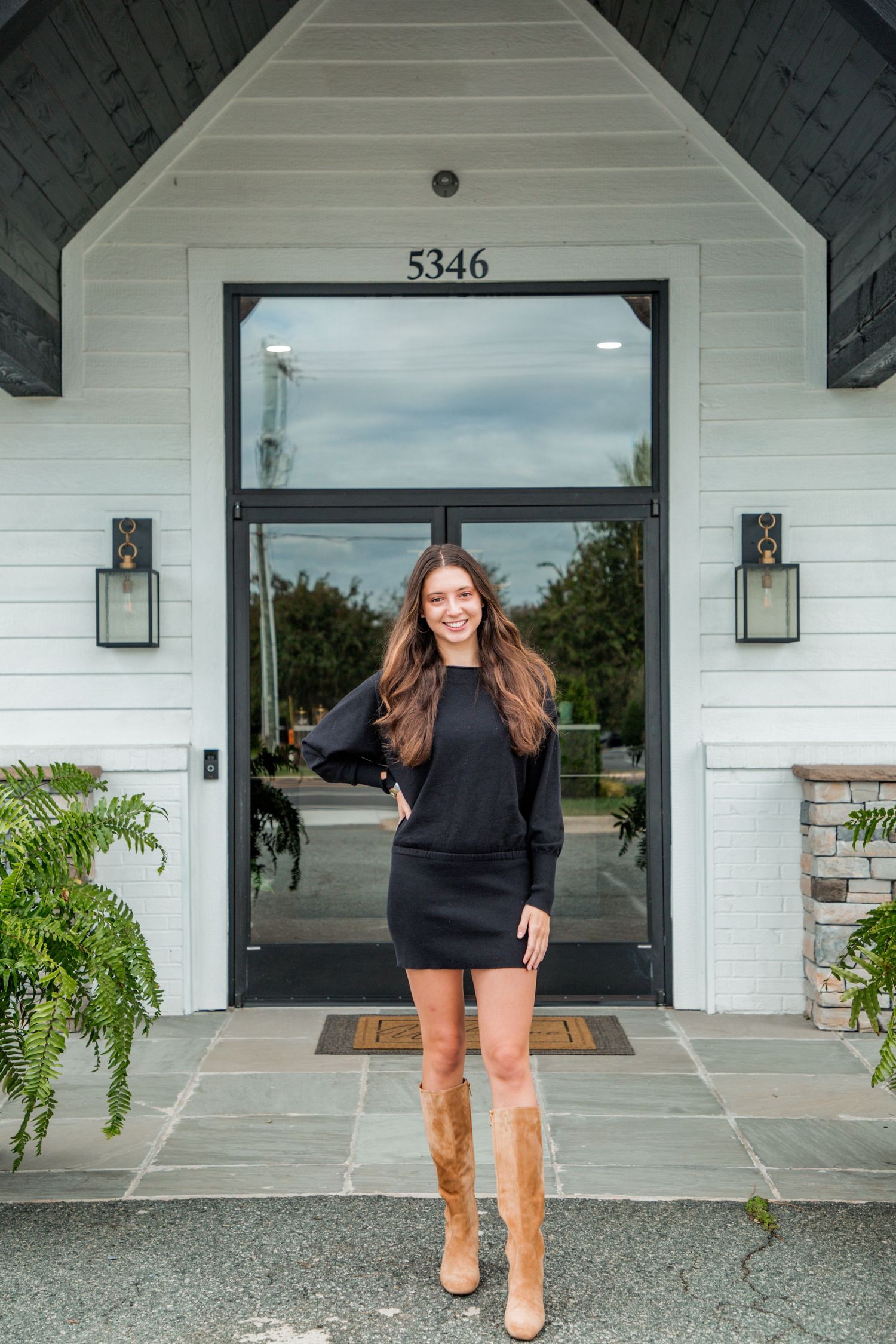 Smiling young woman in black mini dress and tan boots stands before white building entrance 5346.