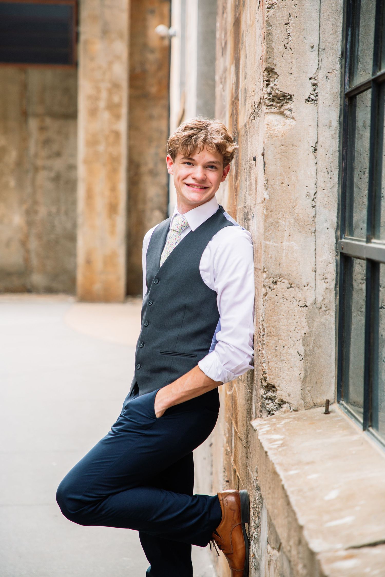 Smiling young man in gray vest white shirt leaning against stone wall beside window outdoors.