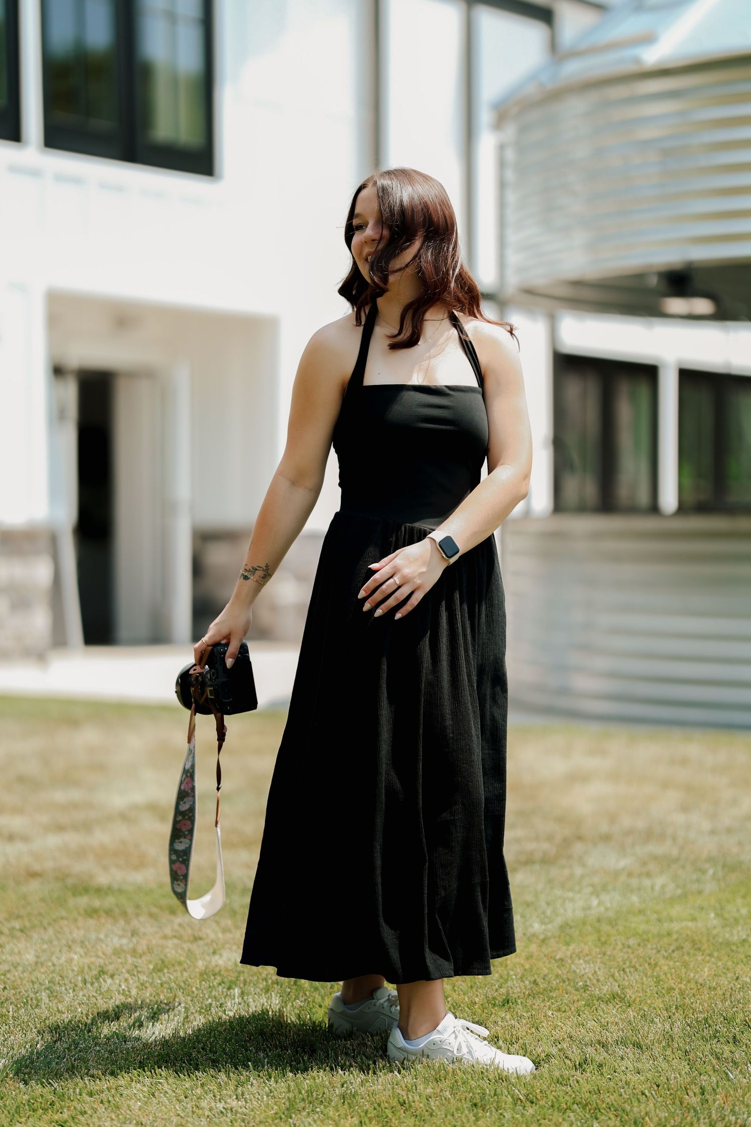 Brunette woman in black halter maxi dress holds camera on green lawn in bright sunlight.