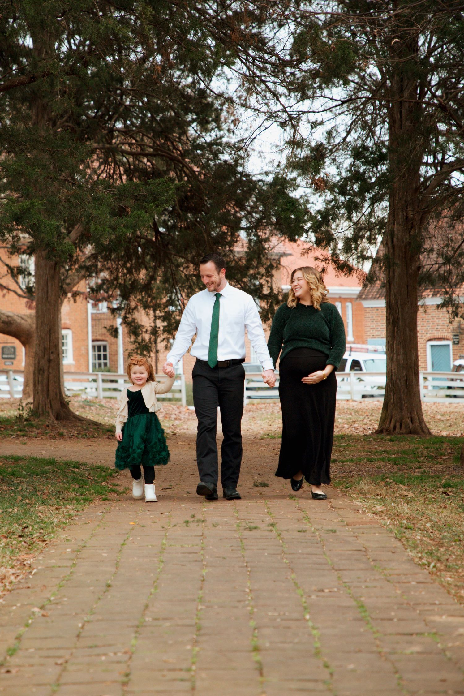 Pregnant mother and father walking with toddler daughter on brick path during maternity photos.