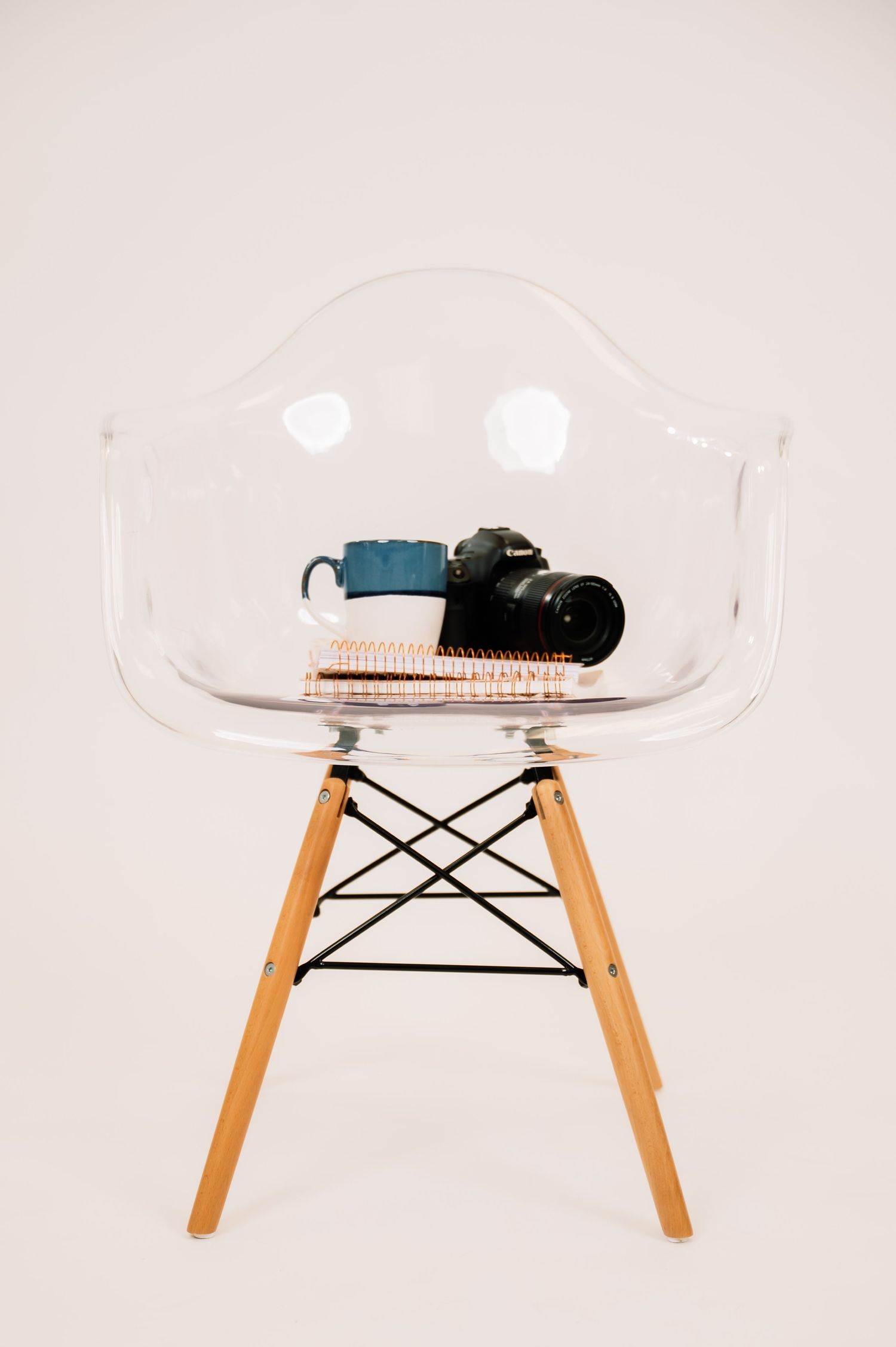 Clear acrylic Eames-style chair with wooden legs holding a blue mug, notebook, and DSLR camera.